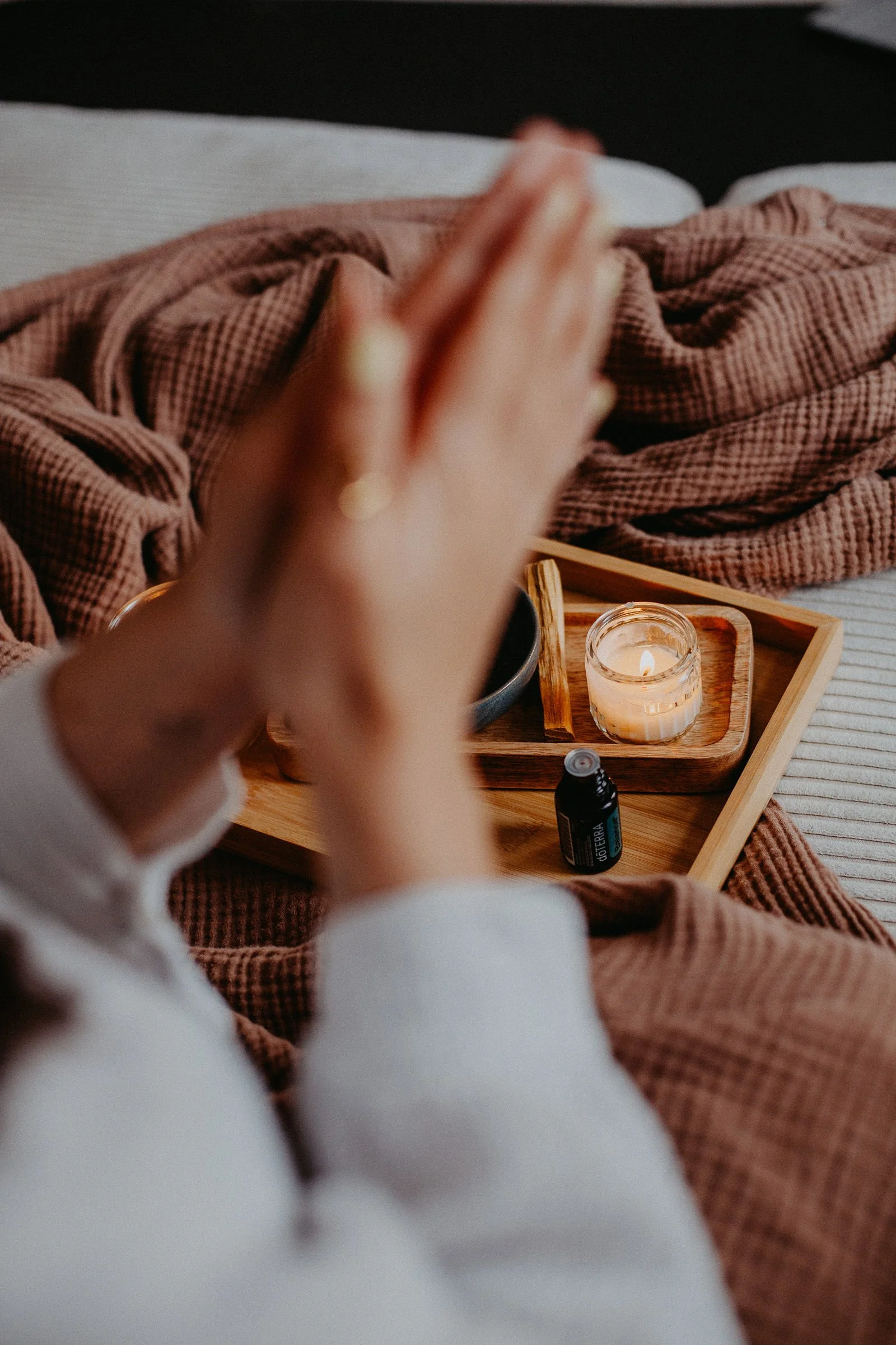 Eine Person sitzt im Bett mit verschränkten Händen, vor einem Holztablett mit einer Kerze, ätherischem Öl und einem Löffel. Das Bett ist mit einer braunen Decke bedeckt.