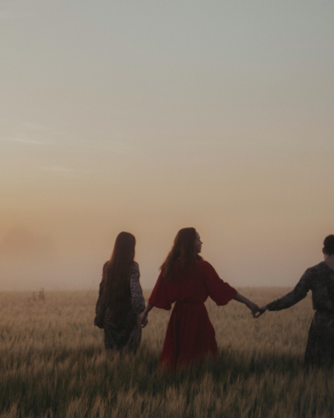 Drei Frauen halten sich an den Händen, spazieren durch ein weites Feld bei Sonnenuntergang.