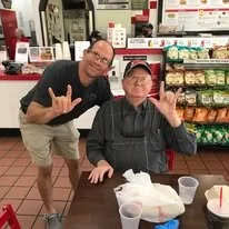 Two men posing inside a store, making hand gestures. One man is standing and smiling, while the other is sitting at a table with food and drinks.
