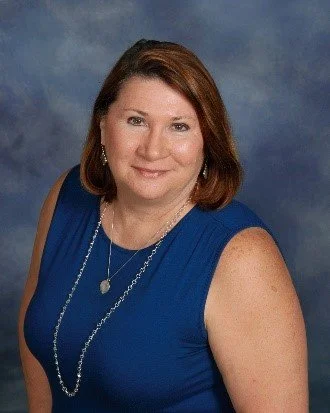 A woman with shoulder-length brown hair wearing a blue sleeveless top, posing against a cloudy blue background.