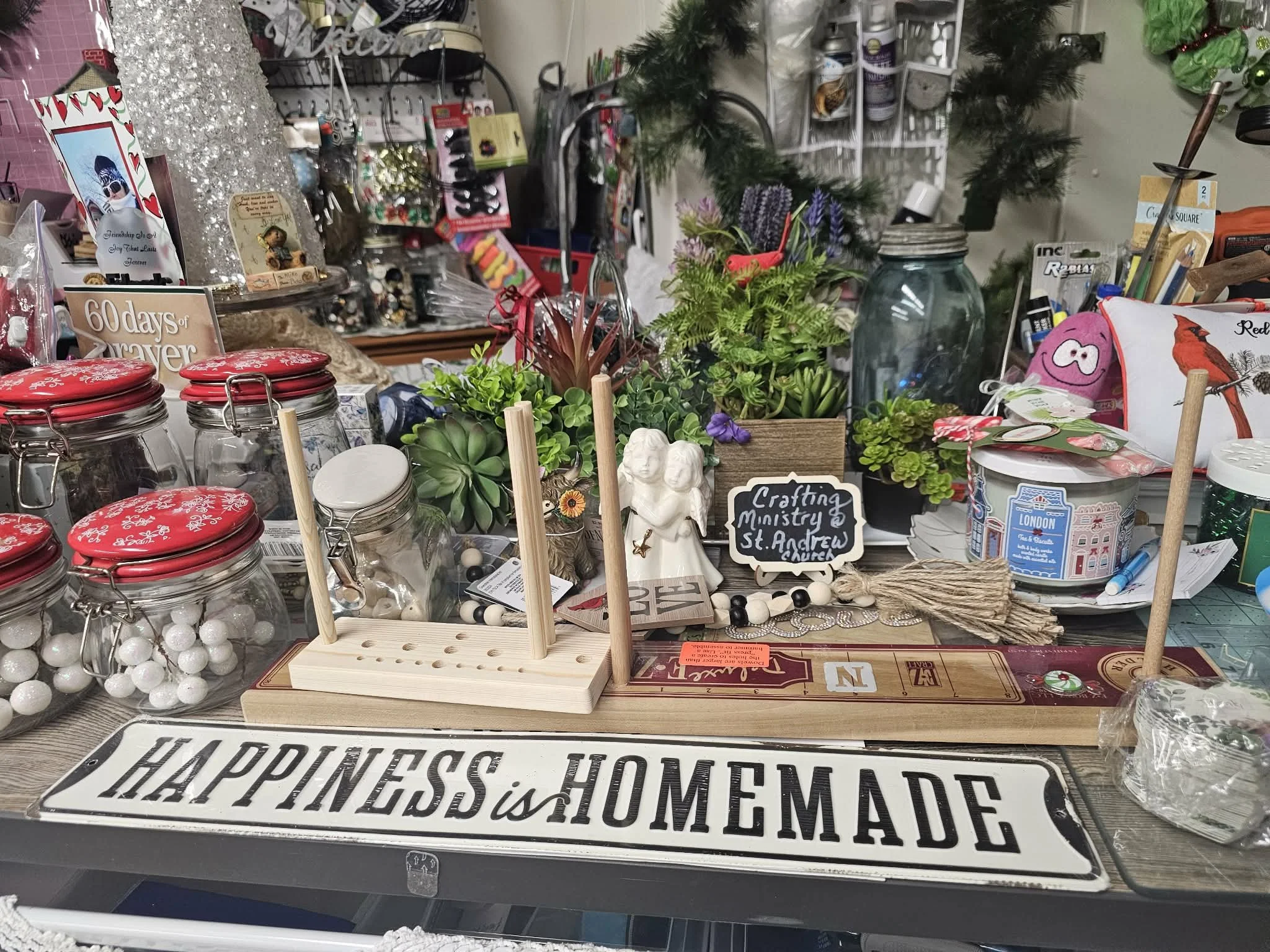 A cluttered craft or thrift store display with various decorative items, plants, jars, and signs. A prominent sign reads 'Happiness is Homemade.'