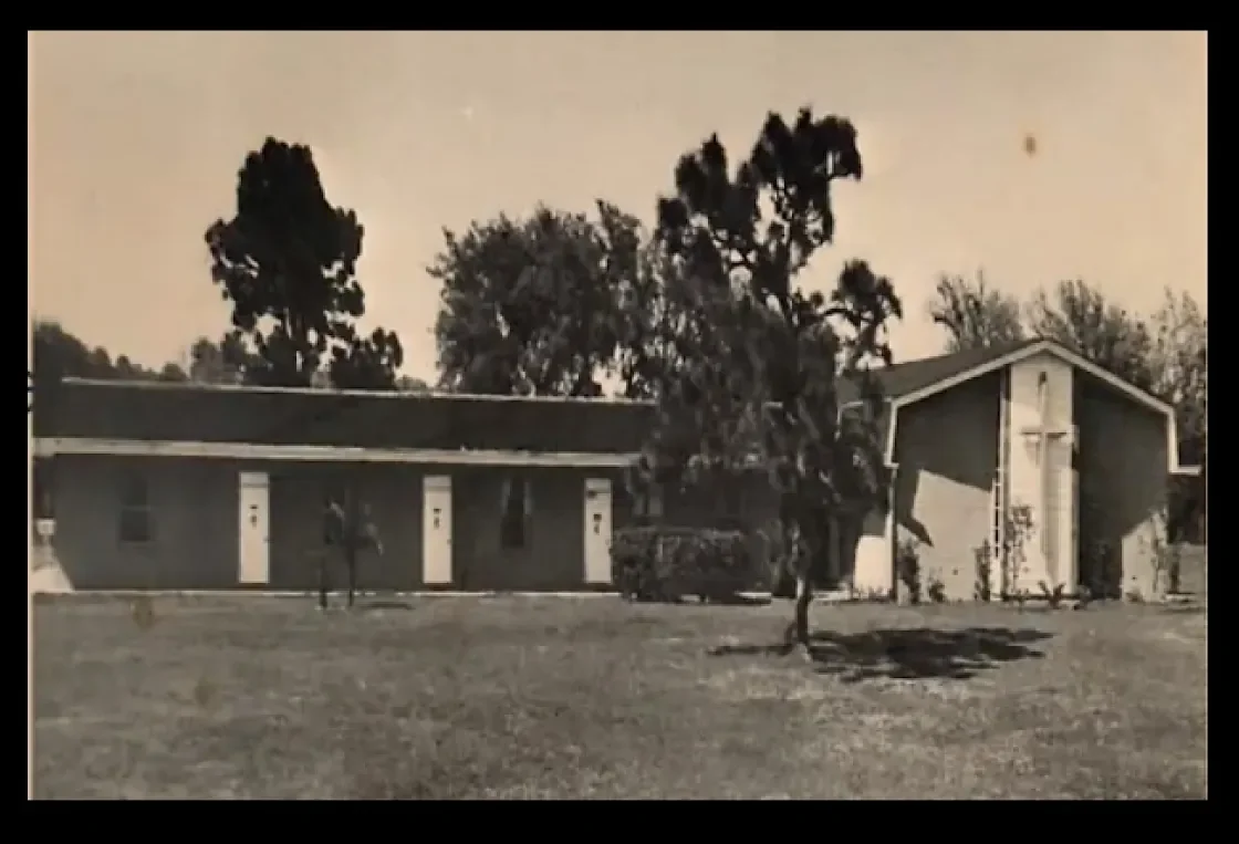 A black and white photo of a church with a cross on the front, surrounded by trees and a grassy area.