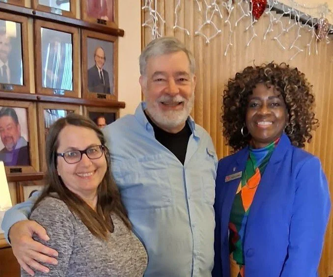 Three smiling people standing close together indoors, with framed photos on the wall behind them and holiday decorations.