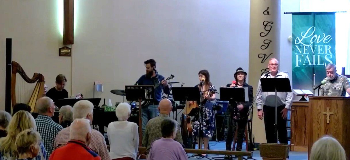 Church service with band and congregation, banner reading "Love Never Fails"