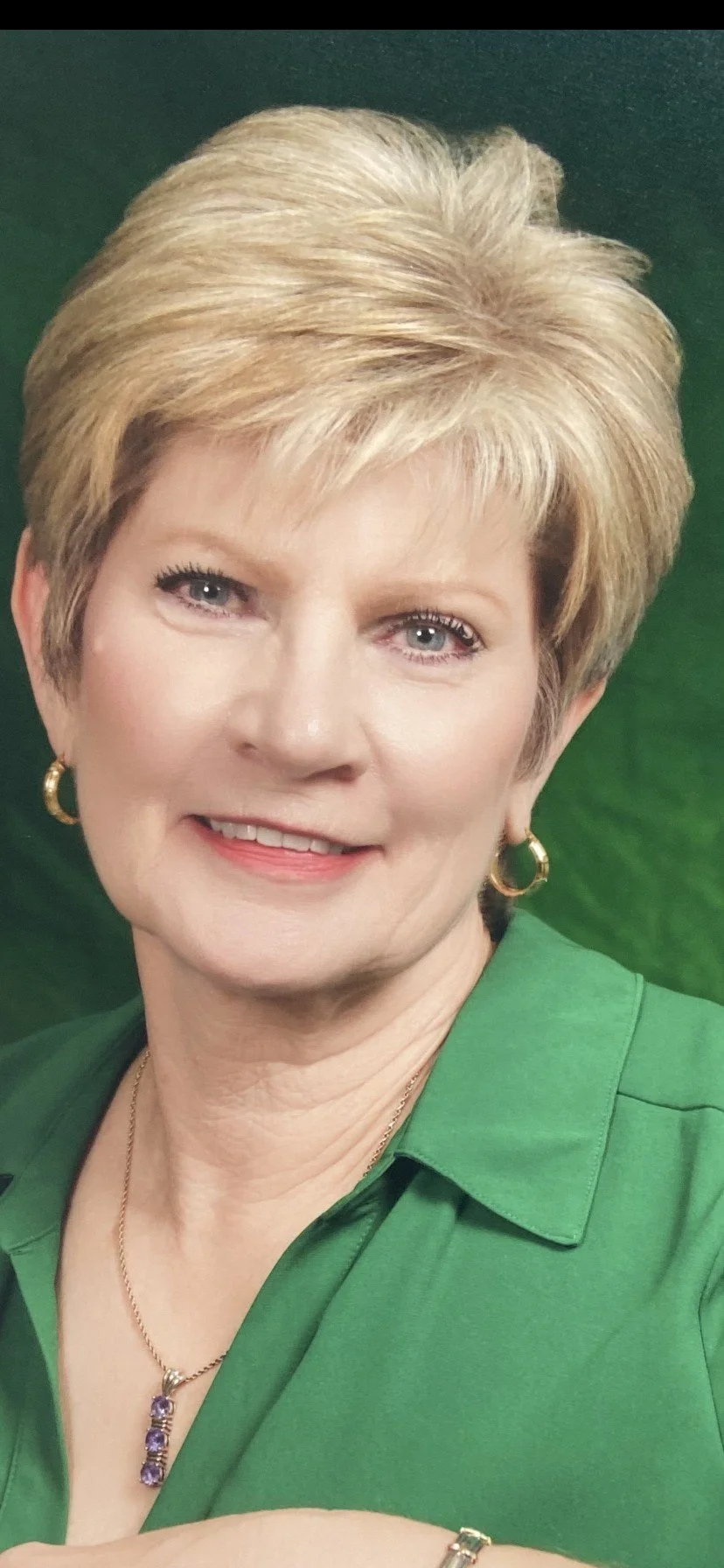 Close-up photograph of a smiling middle-aged woman with short blond hair, wearing gold hoop earrings, a green blouse, and a gold necklace with purple stones against a dark green background.