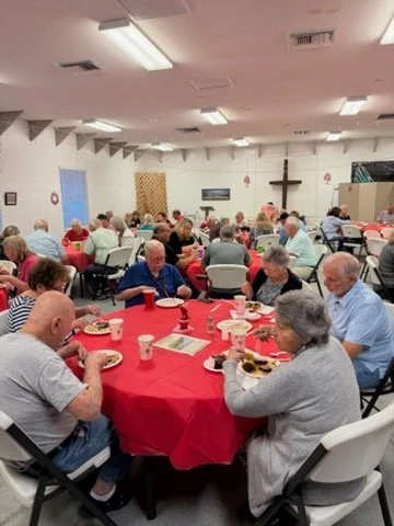 People gathered at tables with red tablecloths in a room with a cross on the wall, likely for a religious or community event.