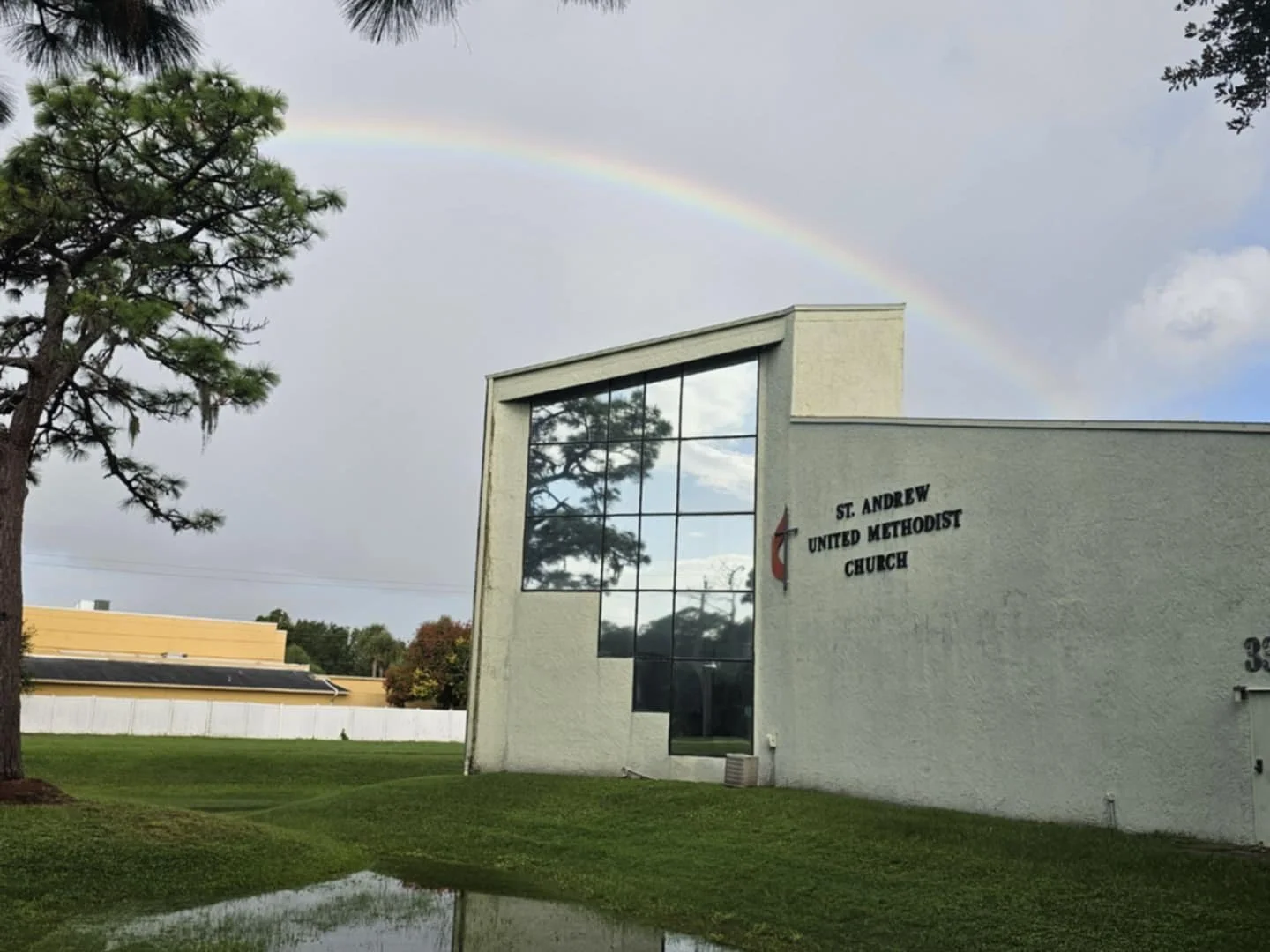 A modern church building with large glass windows and white exterior walls, with a rainbow in the sky above, a tree on the left, and a grassy area in front.