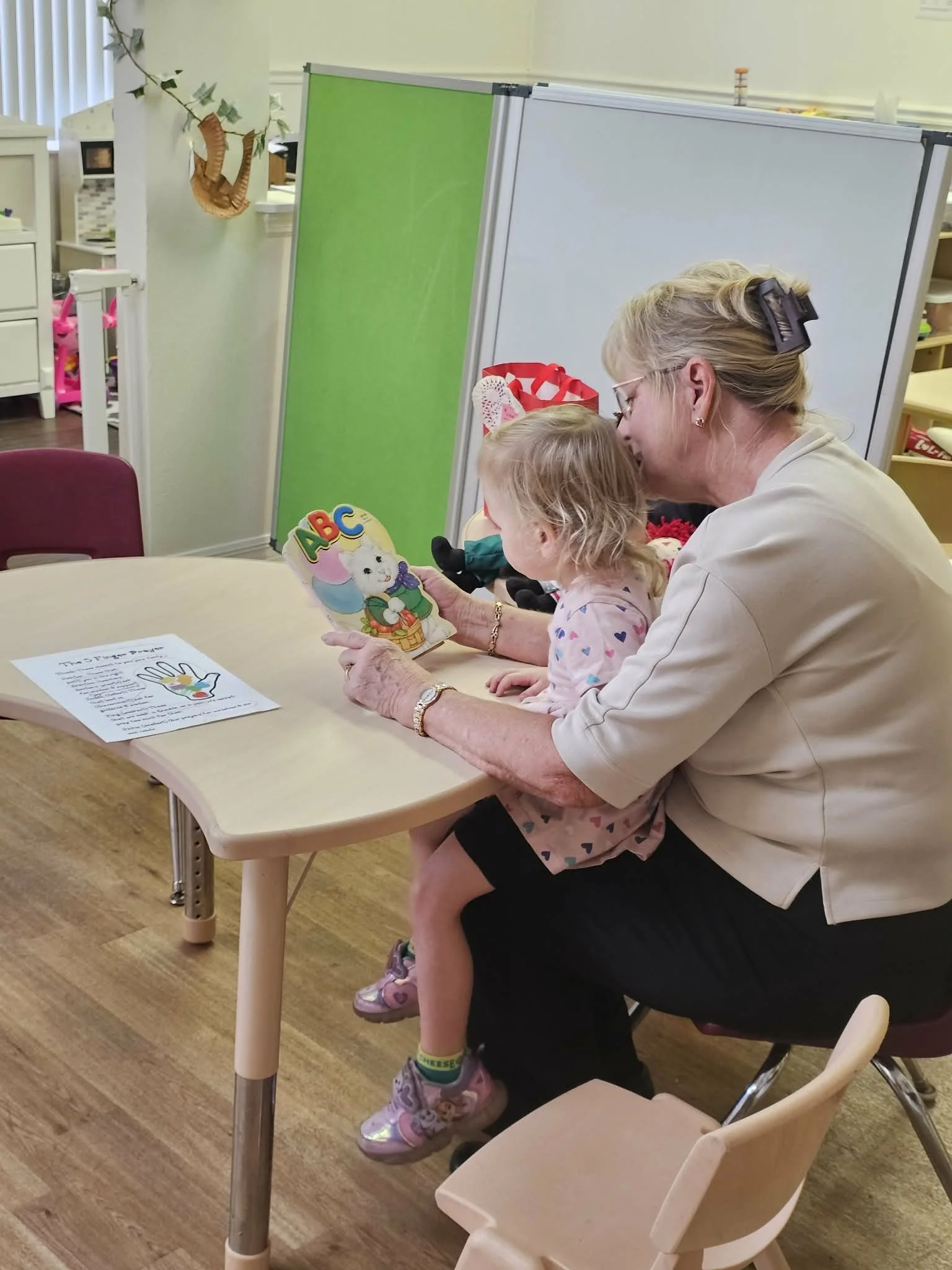 An elderly woman and young girl sit together at a table in a classroom, looking at a colorful book with this book displayed. The classroom has white and greenboards and children's toys and materials in the background.