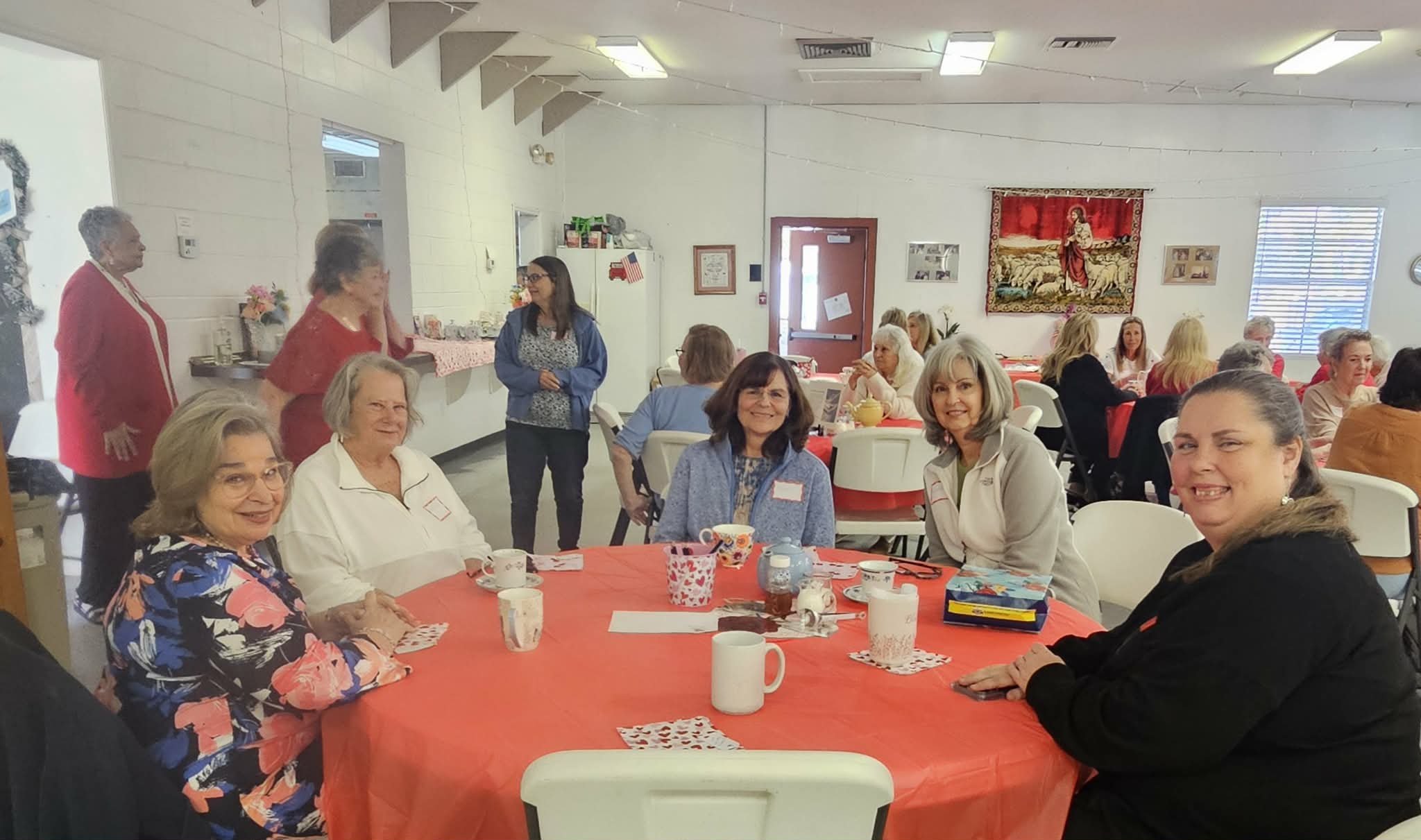 Group of women sitting at a round table with a red tablecloth, smiling and enjoying a social gathering in a decorated room.