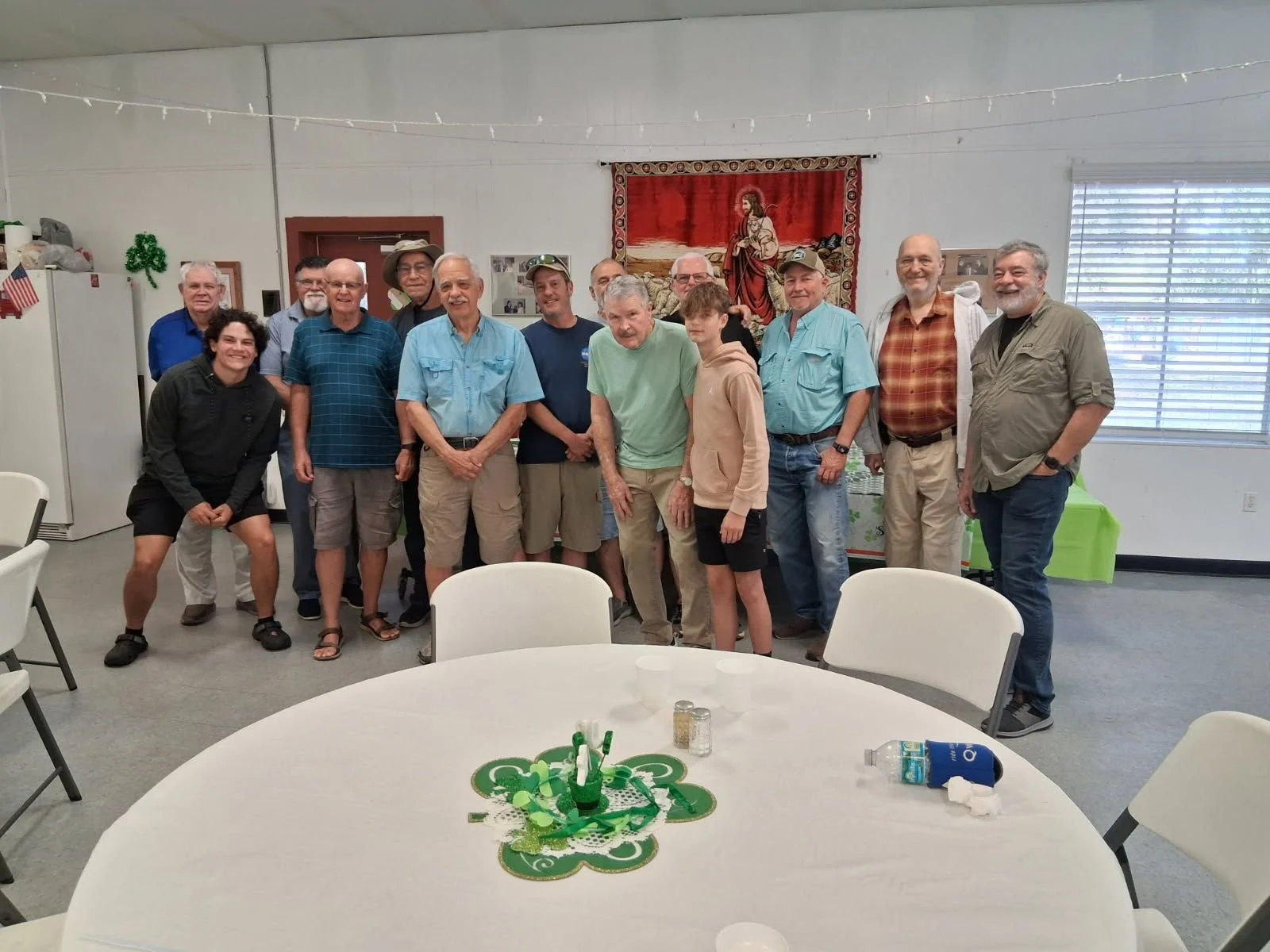 A group of fifteen people gathered in a room decorated for St. Patrick's Day, posing for a photo behind a table with green decorations and a tablecloth with shamrocks. The group includes men of various ages and two boys. There are festive decorations, including a shamrock on the wall, and a religious tapestry in the background. The room has a window with blinds, white walls, and a few chairs and tables in the foreground.
