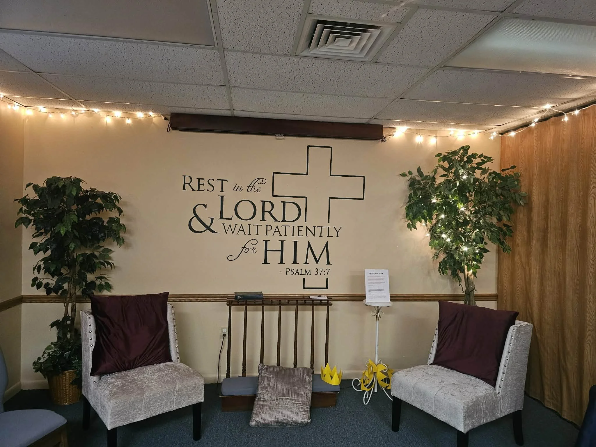 Interior of a room with two beige chairs with purple cushions, a small table in between with yellow and gray decorative items, potted plants on either side, string lights on the ceiling, a Christ-like cross drawing on the wall with Bible verse Psalm 37:7, and a wooden wall panel on the right.
