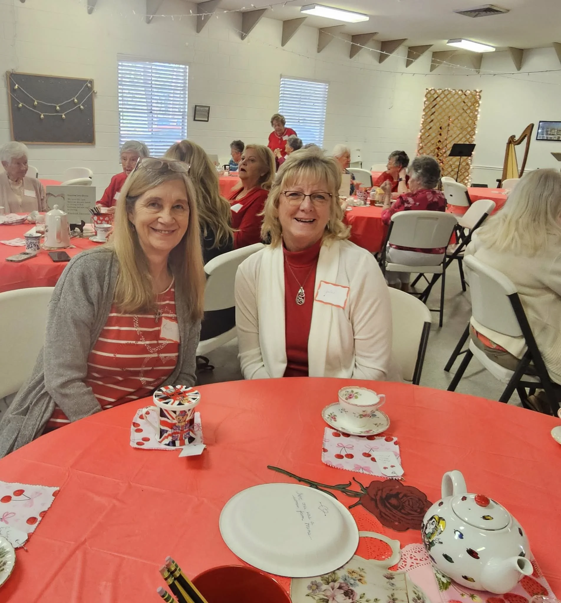 Two women seated at a table during a festive gathering, with several other women in the background, all in a decorated event space with red and white themed decorations.