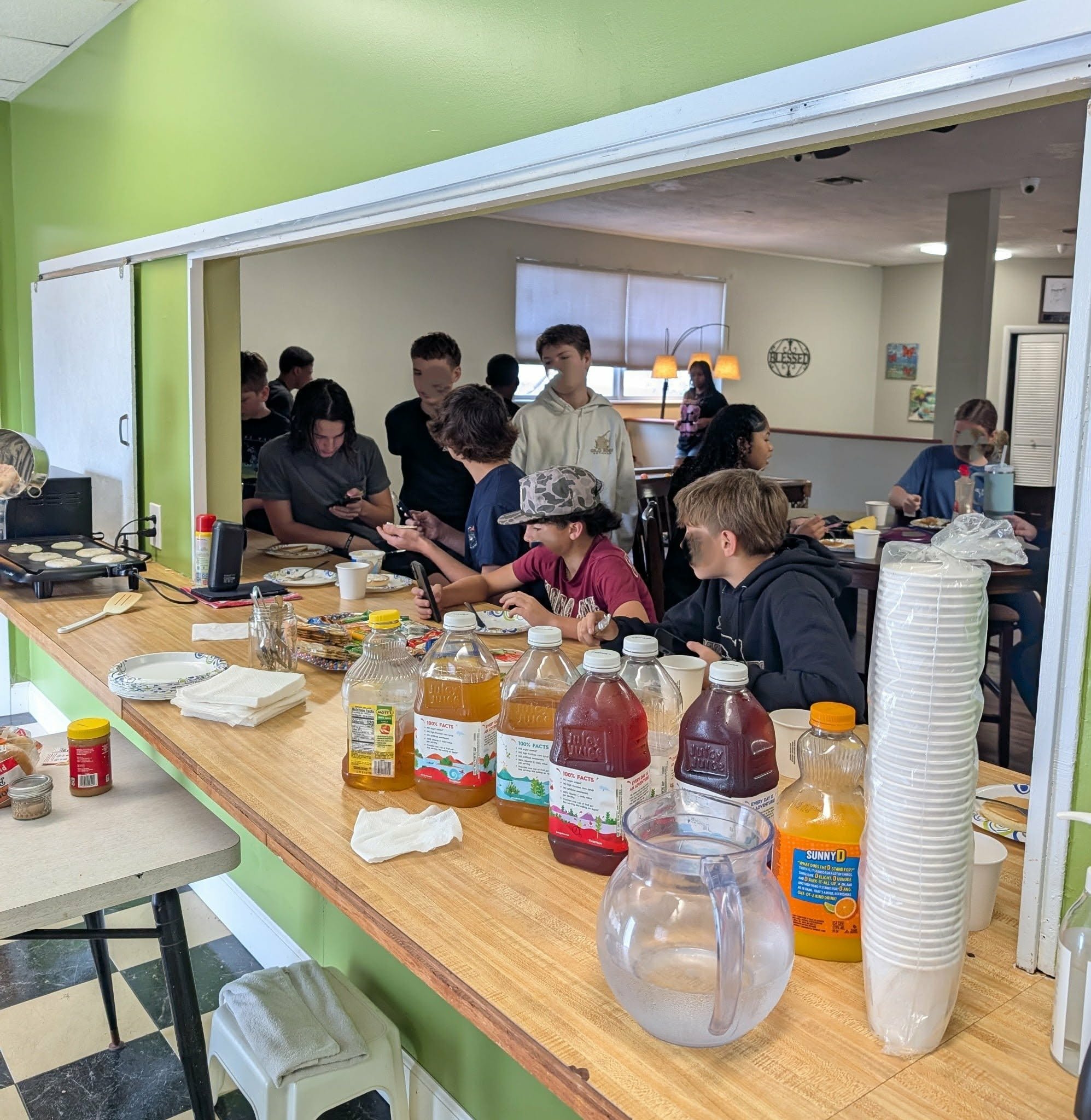 Group of children and teenagers at a breakfast buffet in a kitchen or dining area, with various drinks, plates, and snacks on the counter.
