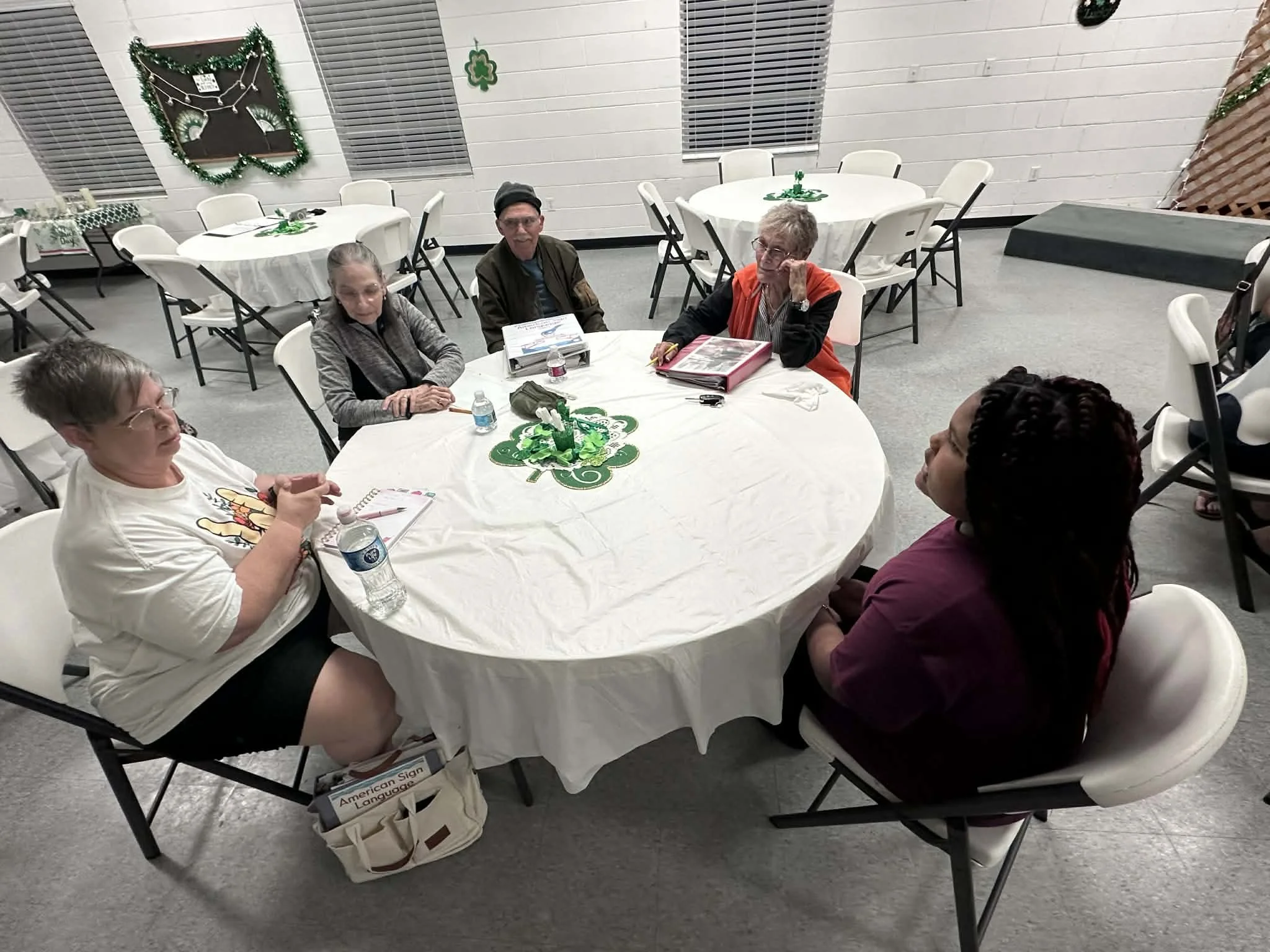 Six people sitting around a table with a St. Patrick's Day decorations in a room with white brick walls and shuttered windows.