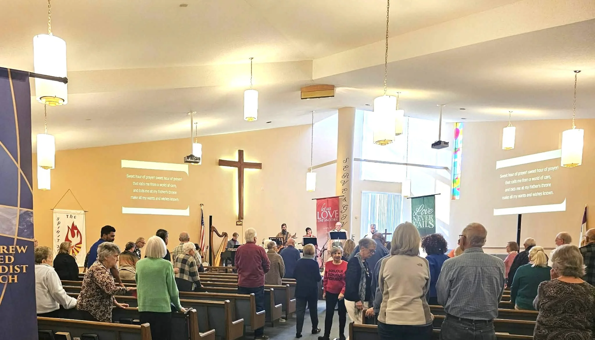 People gathered inside a church for service, standing and facing the front where a band and choir are performing. There is a large cross on the wall behind them, with hymn lyrics projected on the wall.