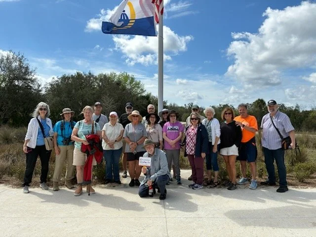 Group of people standing outdoors on a sunny day, with a flagpole and partial American flag visible in the background.