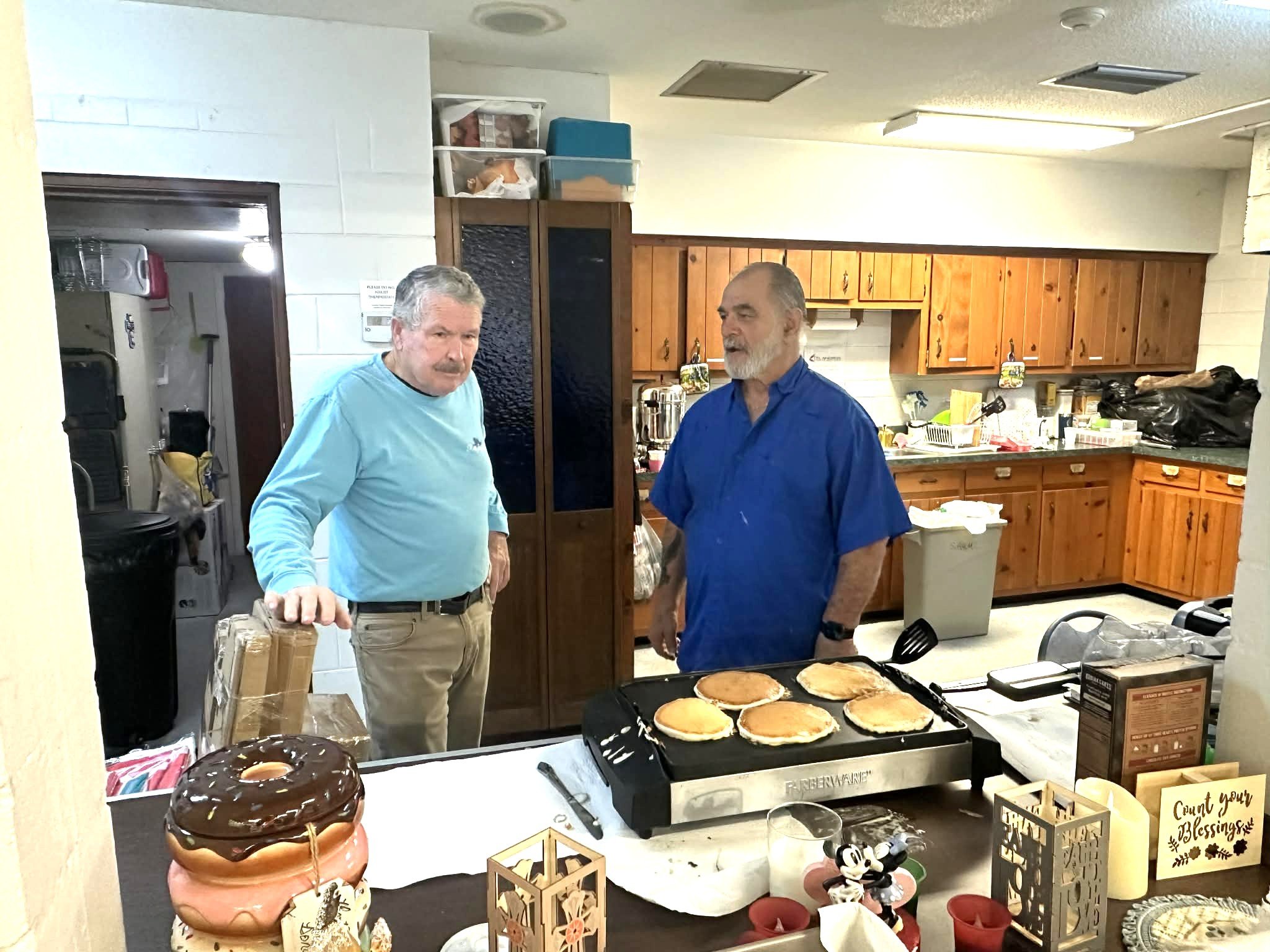 Two men in a kitchen standing near a griddle with four pancakes cooking, one man in a blue shirt and the other in a light blue shirt, surrounded by kitchen items and holiday decorations.