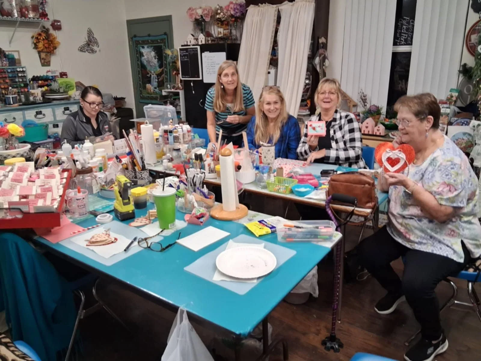 Women and girls sitting around a craft table with supplies, smiling and holding Valentine-themed decorations in a craft room.