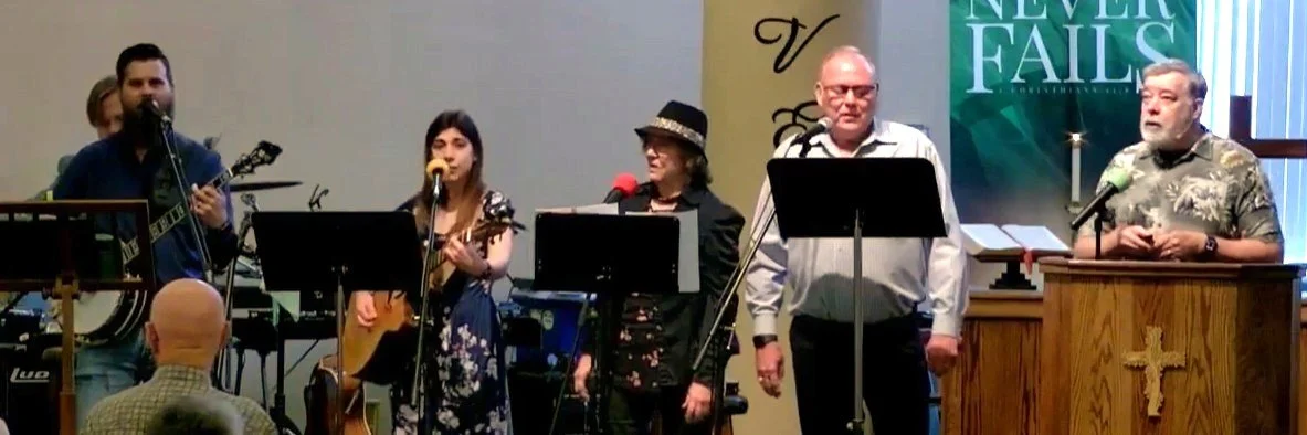 A group of six people singing and playing instruments on stage during a church service. There are microphones and music stands, and a banner reading 'Never Fails' is visible in the background. One person is standing behind a wooden podium with a cross symbol.