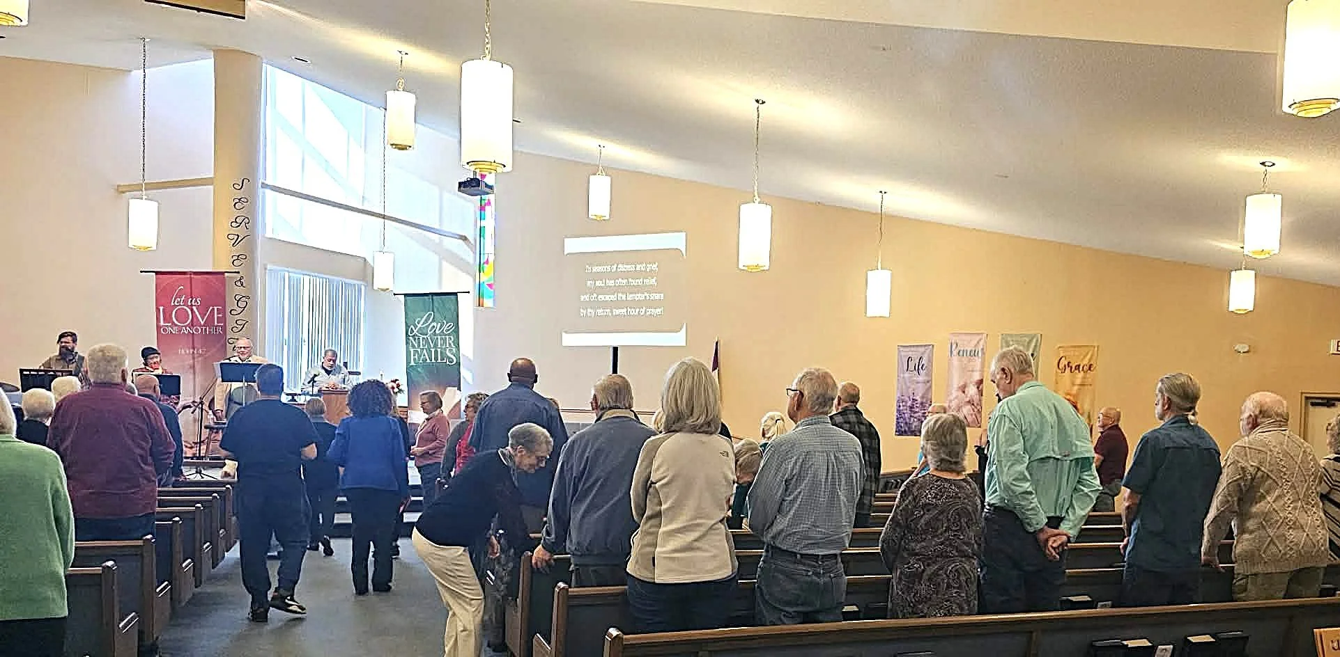 People gathering in a church sanctuary during a service or event, some standing and some seated in pews, with banners hanging on the walls displaying messages like 'Let us love one another,' 'Love never fails,' 'Life,' 'Peace,' and 'Grace,' and a screen displaying lyrics or scripture.