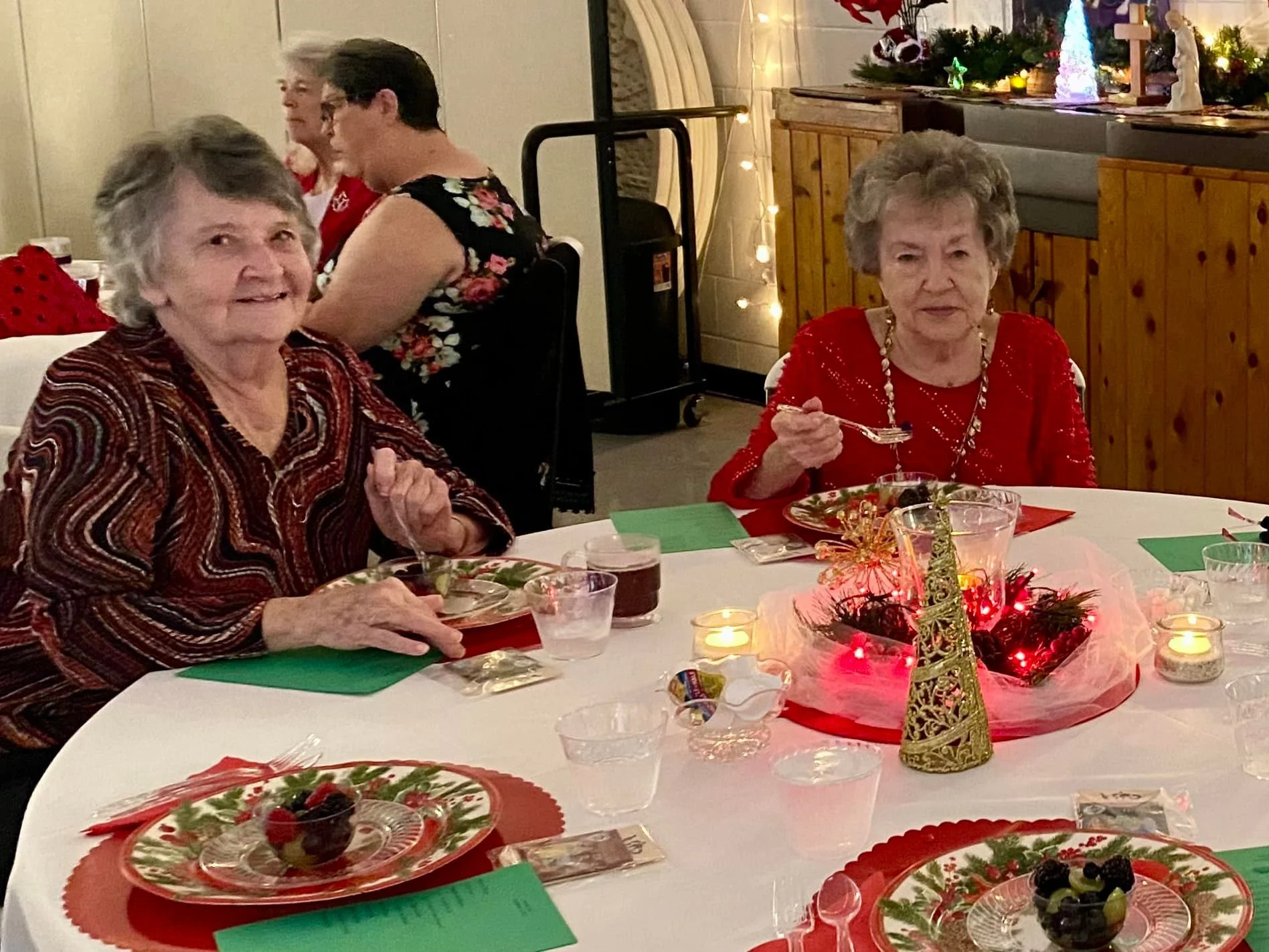 Two elderly women sitting at a decorated Christmas dinner table with festive centerpieces, candles, and plates of fruit, enjoying a holiday meal.