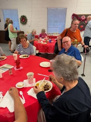 Group of elderly people at a holiday party dining together with Christmas decorations, including a wreath and red garland, in the background.