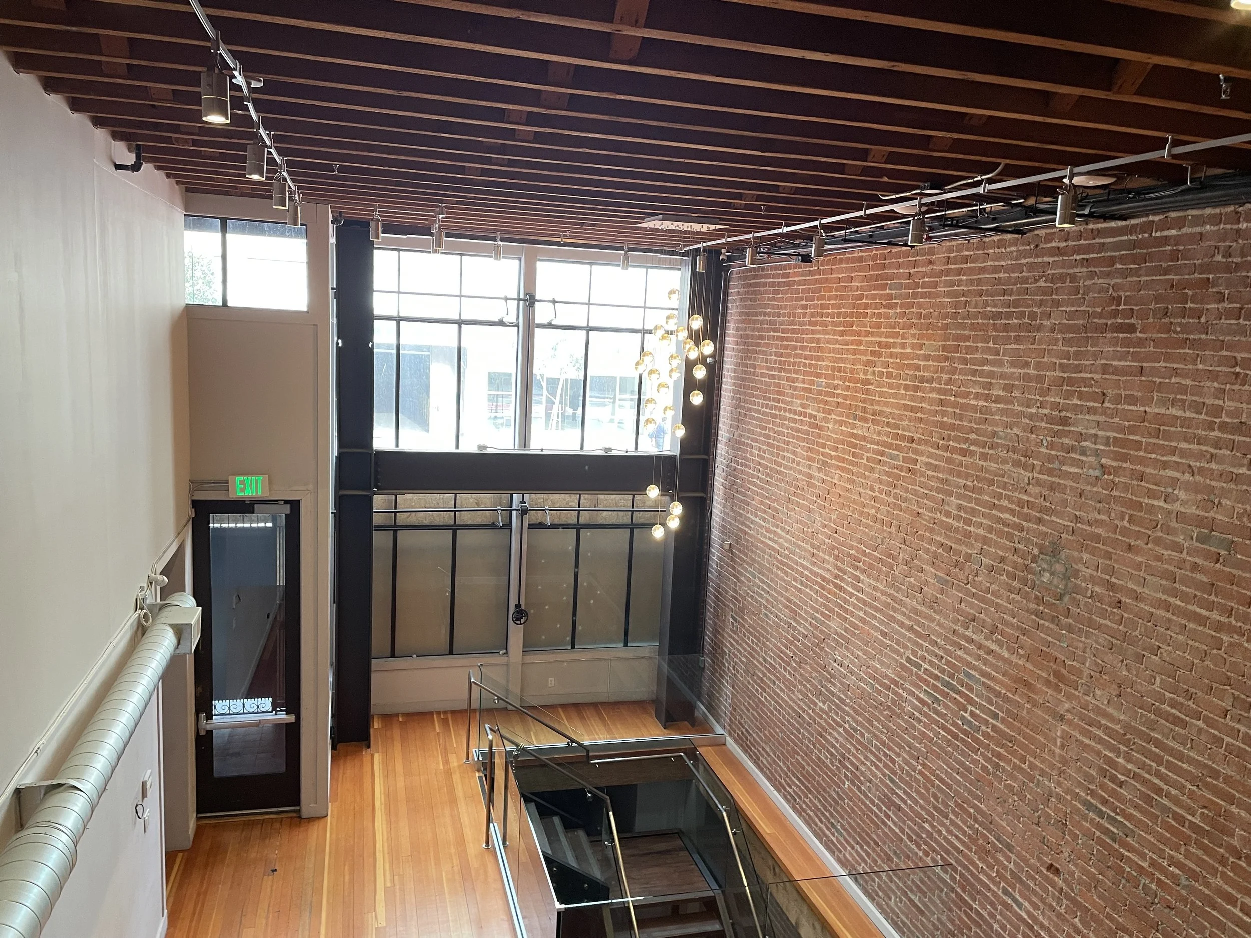 Interior of a modern building with a high ceiling, large front windows, exposed brick wall, wood flooring, and a staircase with glass railings. Structural engineering by BASE Design in San Francisco.