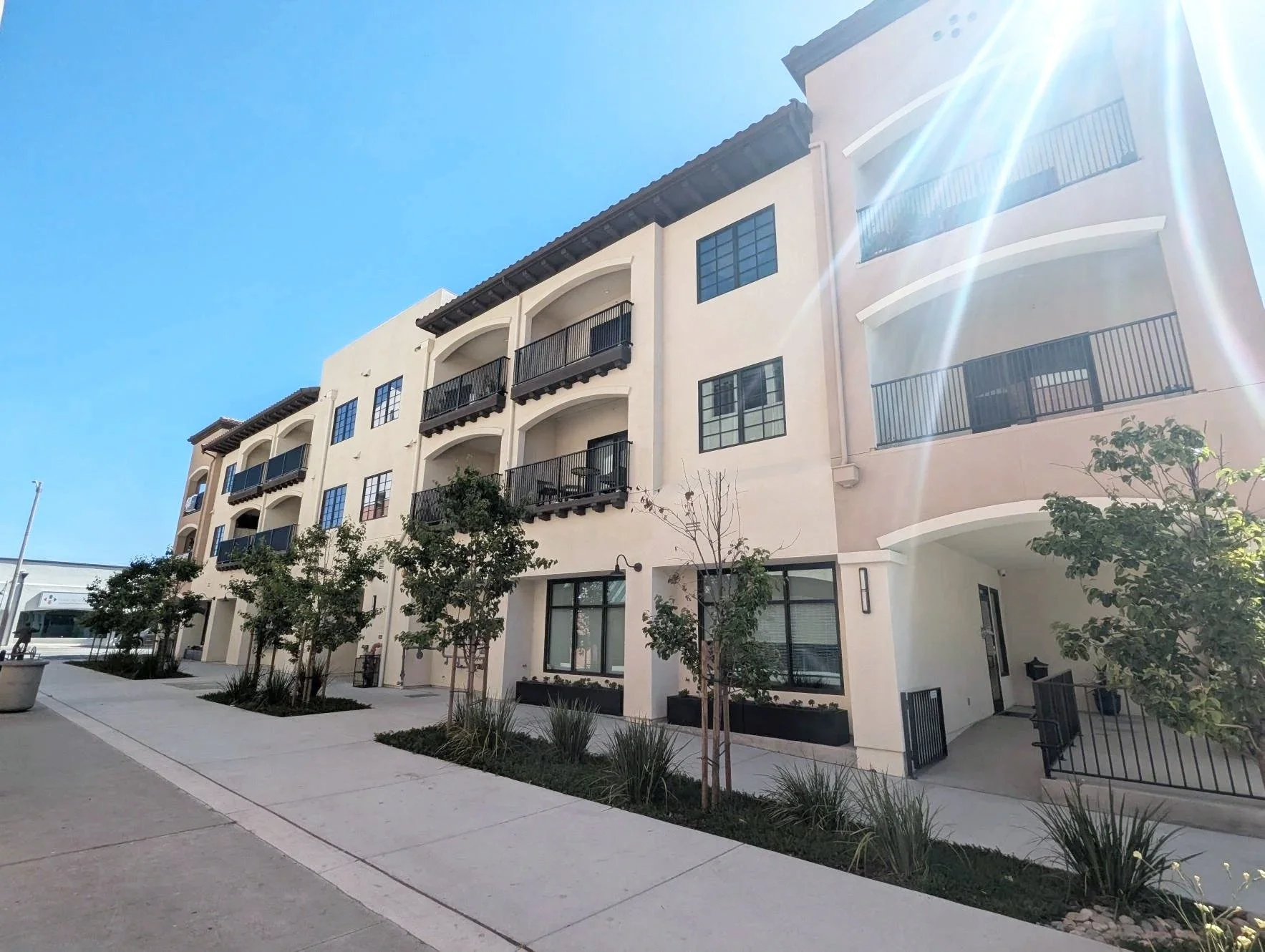 A modern multi-story apartment building with balconies, large windows, and landscaped trees outside on a sunny day. Structural engineering by BASE Design in San Francisco.