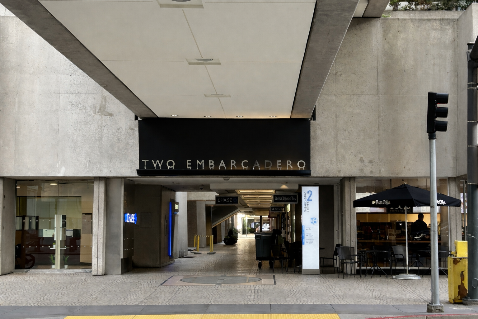 Street view of Two Embarcadero storefront with entrance, outdoor seating, umbrellas, and a traffic light in front.
