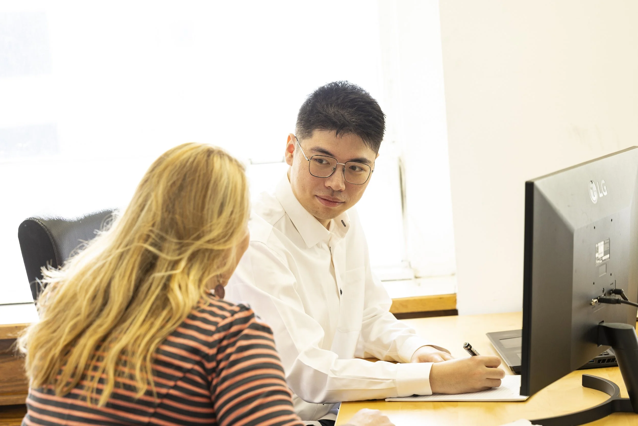 A man from BASE Design Structural Engineering in San Francisco sits with a female client, having a conversation at a desk in an office, with a computer monitor on the desk.
