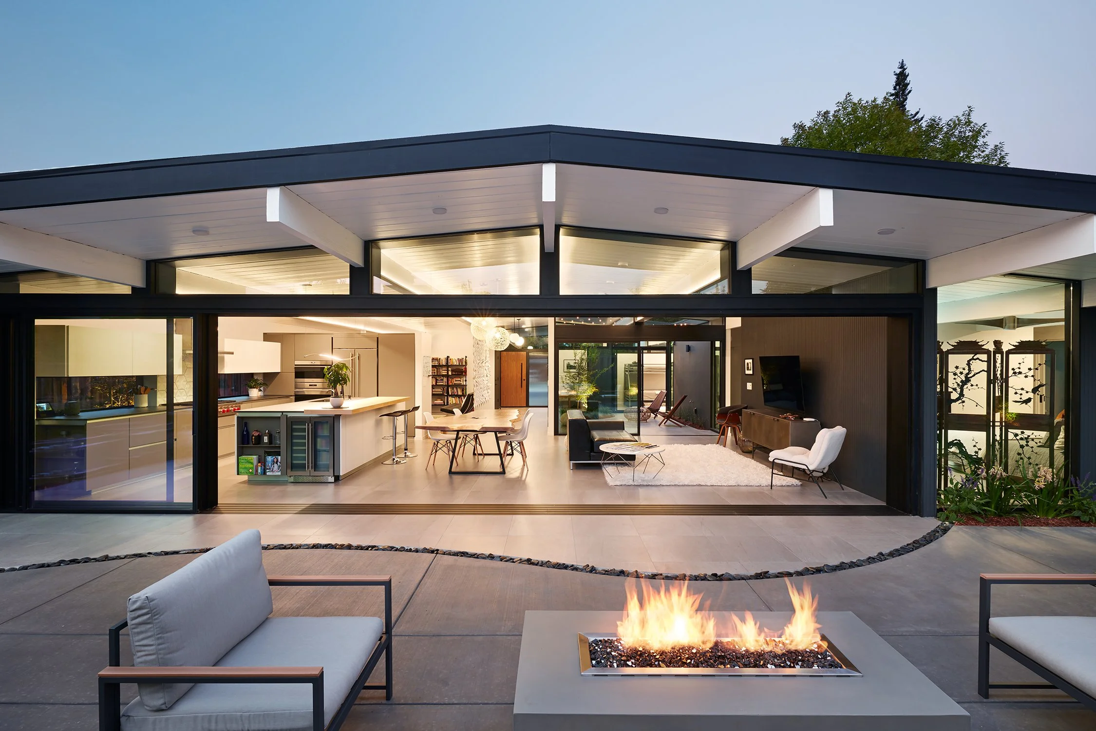 Rear view of an Eichler-designed home in Palo Alto, CA, with large glass sliding doors, outdoor fire pit, and patio furniture, overlooking a well-lit interior living and dining area. Structural engineering by BASE Design in San Francisco.