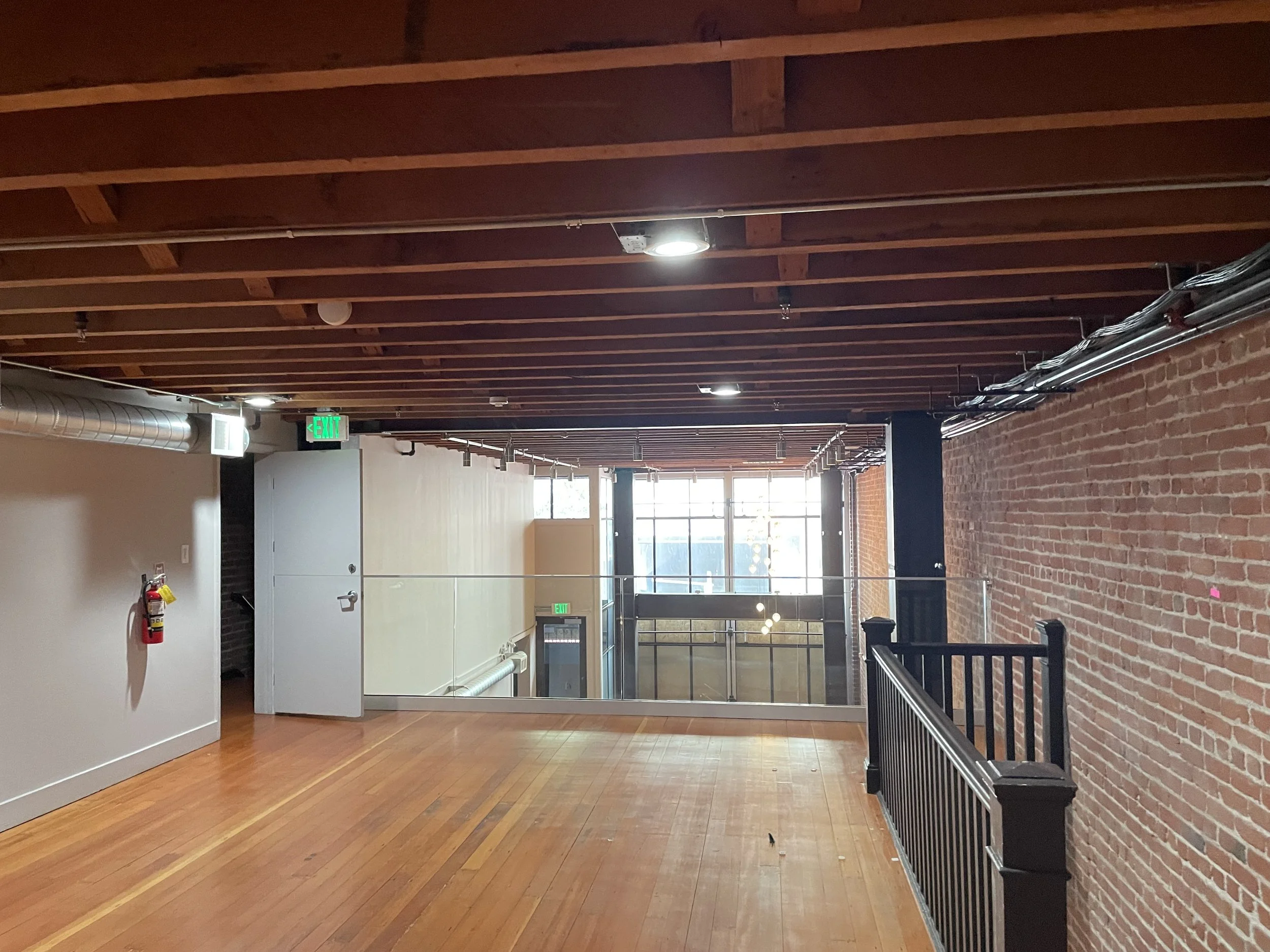 Empty indoor space with wooden floor and exposed brick walls, black railing, windows letting in natural light, ceiling with wooden beams, track lighting, and an exit sign. Structural engineering by BASE Design in San Francisco.