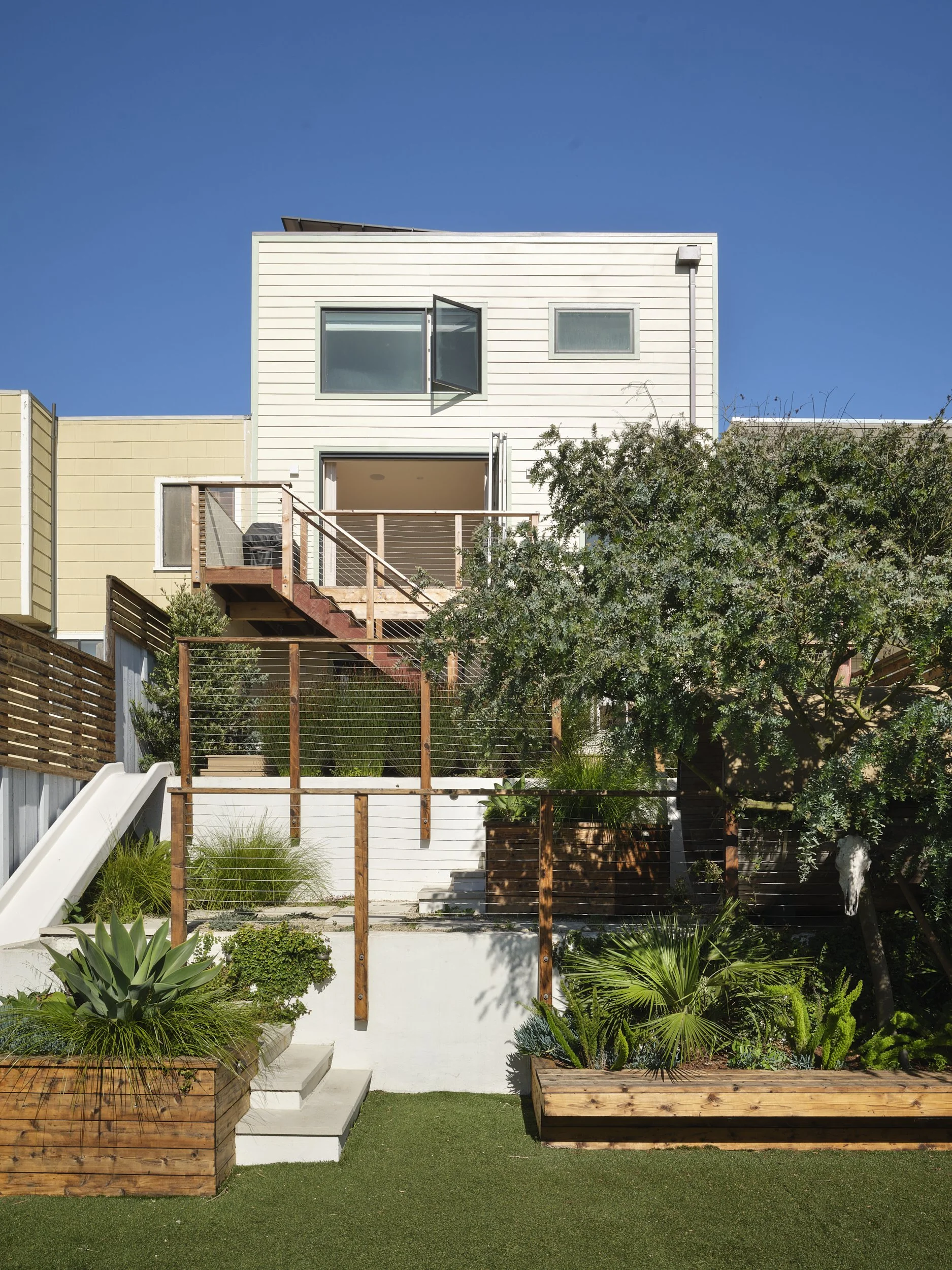 Modern multi-story house with white exterior, open window, small second window, and balcony with railing, situated behind a landscaped yard with plants, trees, and wooden planters, under a clear blue sky. Structural engineering by BASE Design in San