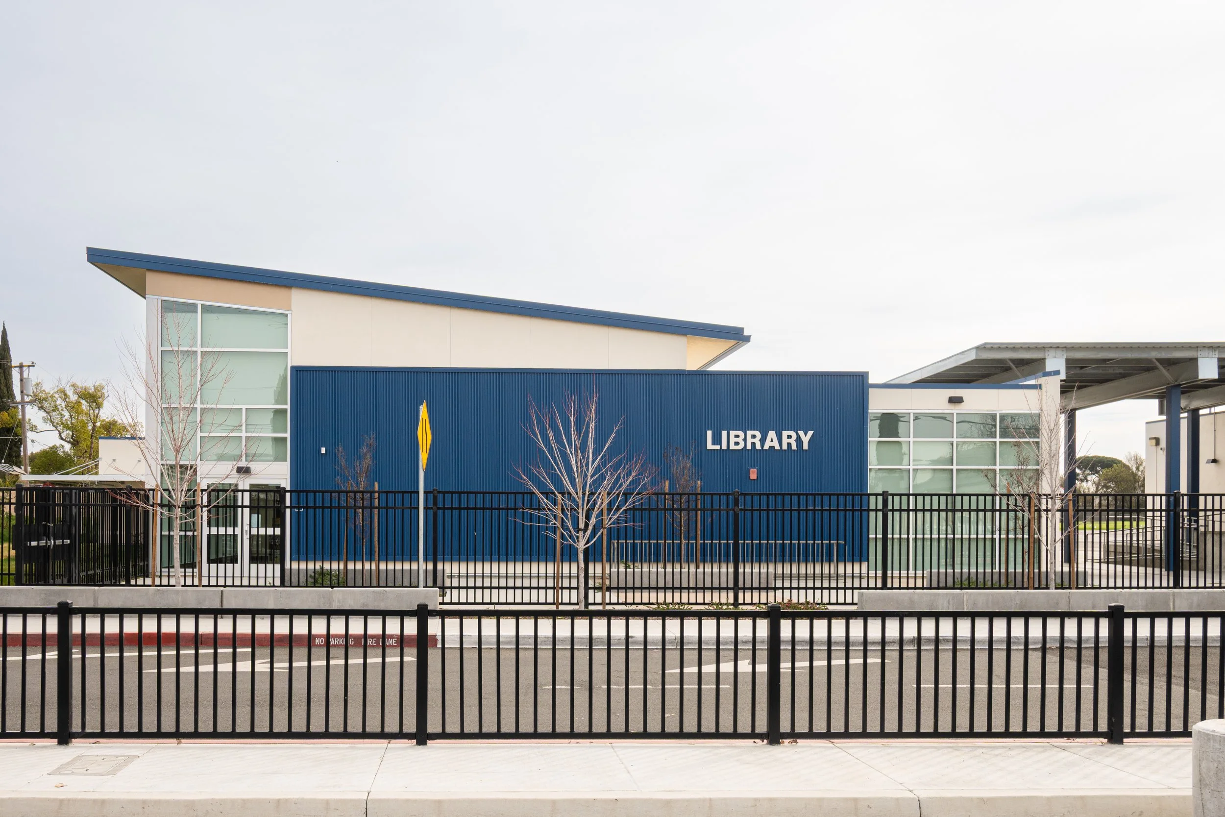 A modern school library building with blue and beige exterior walls, large glass windows, and a sign reading 'LIBRARY'. There is a black metal fence and a sidewalk in front. Structural engineering by BASE Design in San Francisco.
