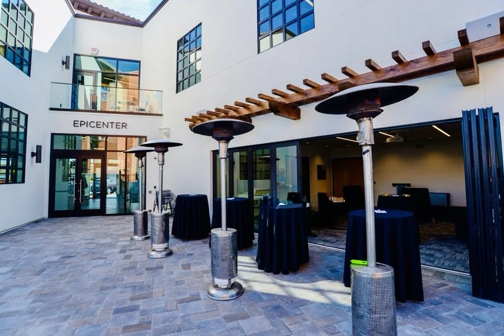 Outdoor patio area with high tables covered in black tablecloths and patio heaters, outside a modern building with large windows and a glass entrance labeled "EPICENTER." Structural engineering by BASE Design in San Francisco.
