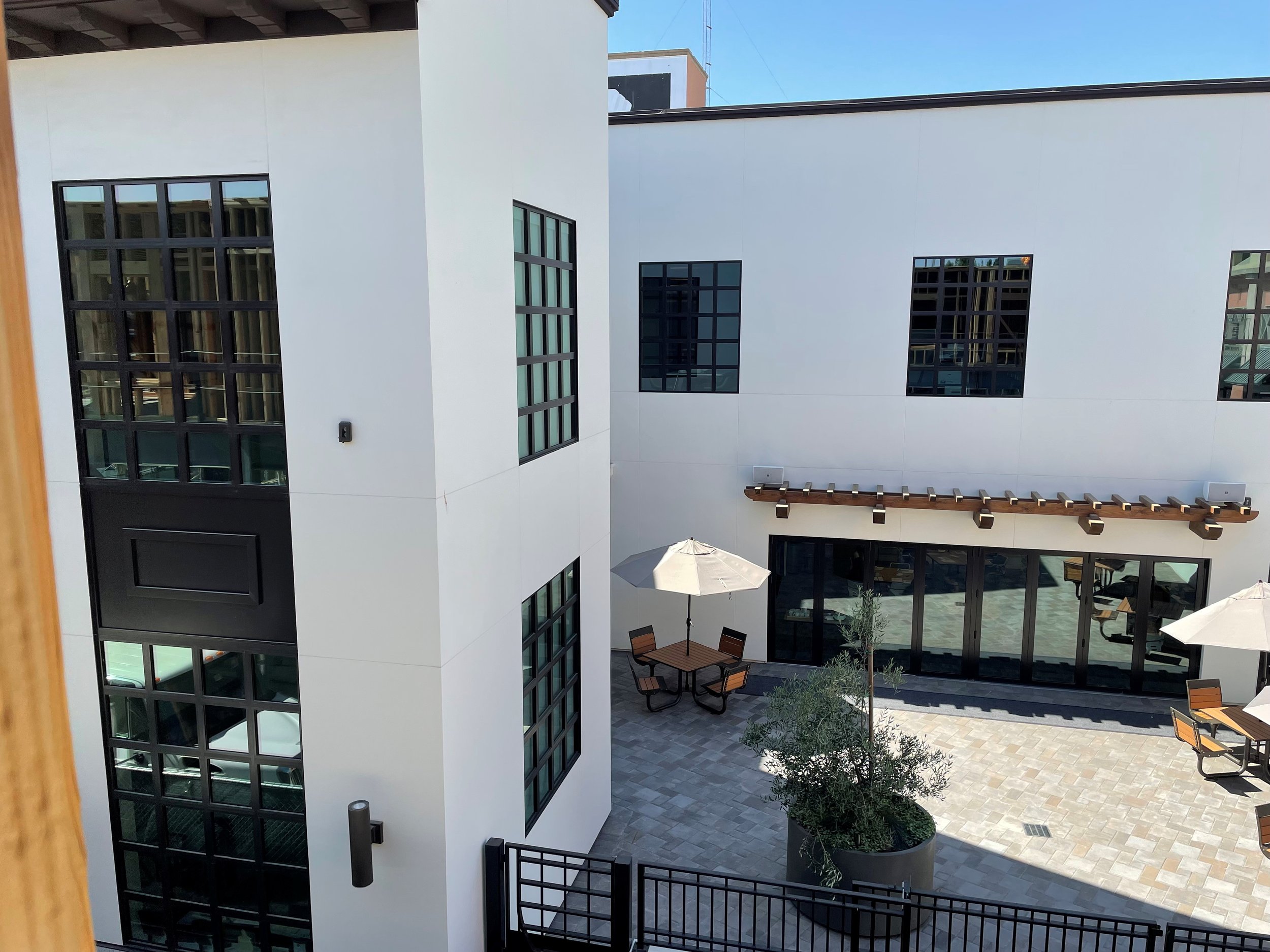 View of a modern building's outdoor courtyard with white walls, black-framed windows, patio tables with umbrellas, and planters with trees. Structural engineering by BASE Design in San Francisco.