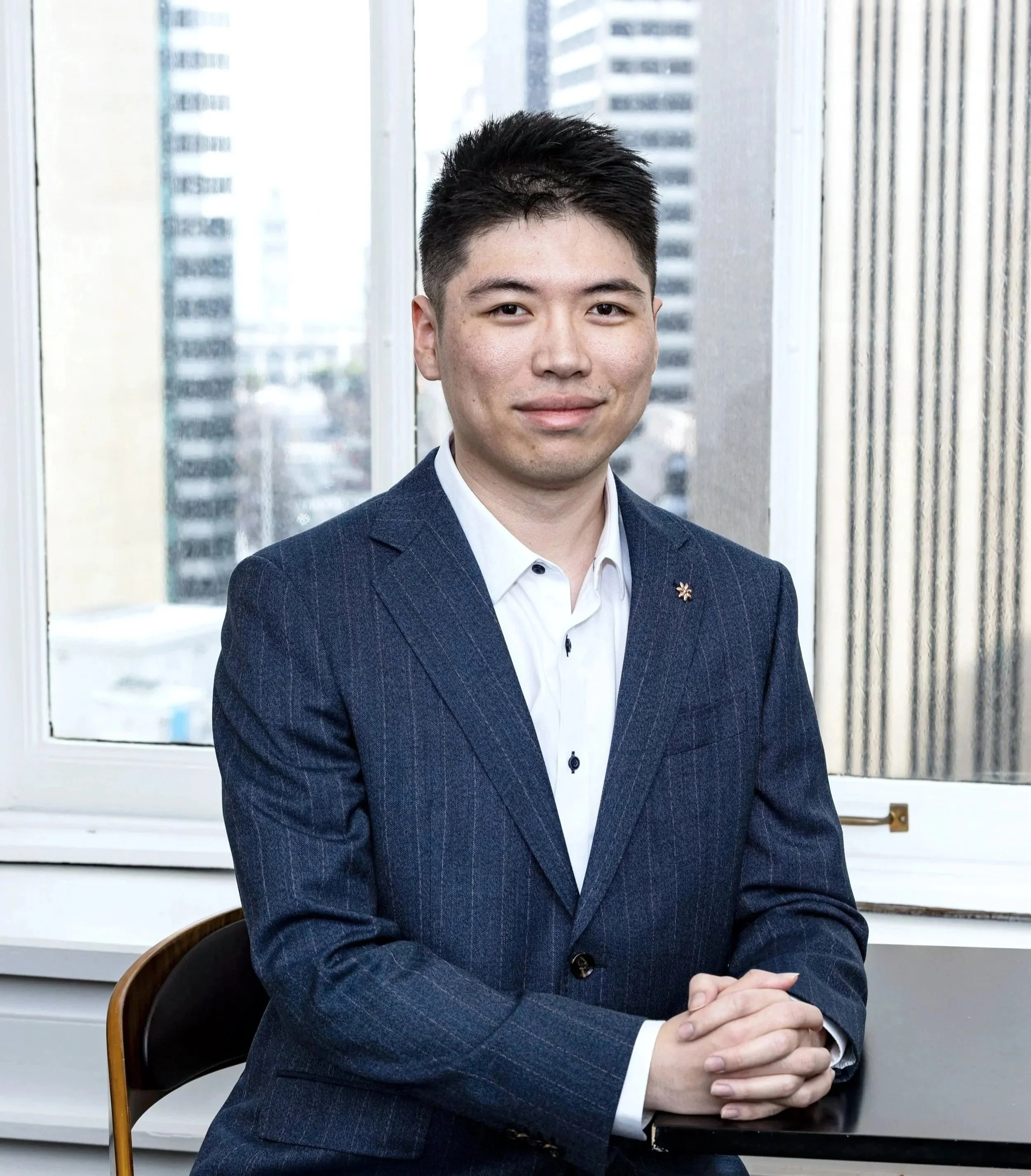 Bosun Liu, a young man in a navy suit and white shirt sitting at a desk in front of a window with city buildings in the background. He's a member of the BASE Design Structural Engineering firm in San Francisco.
