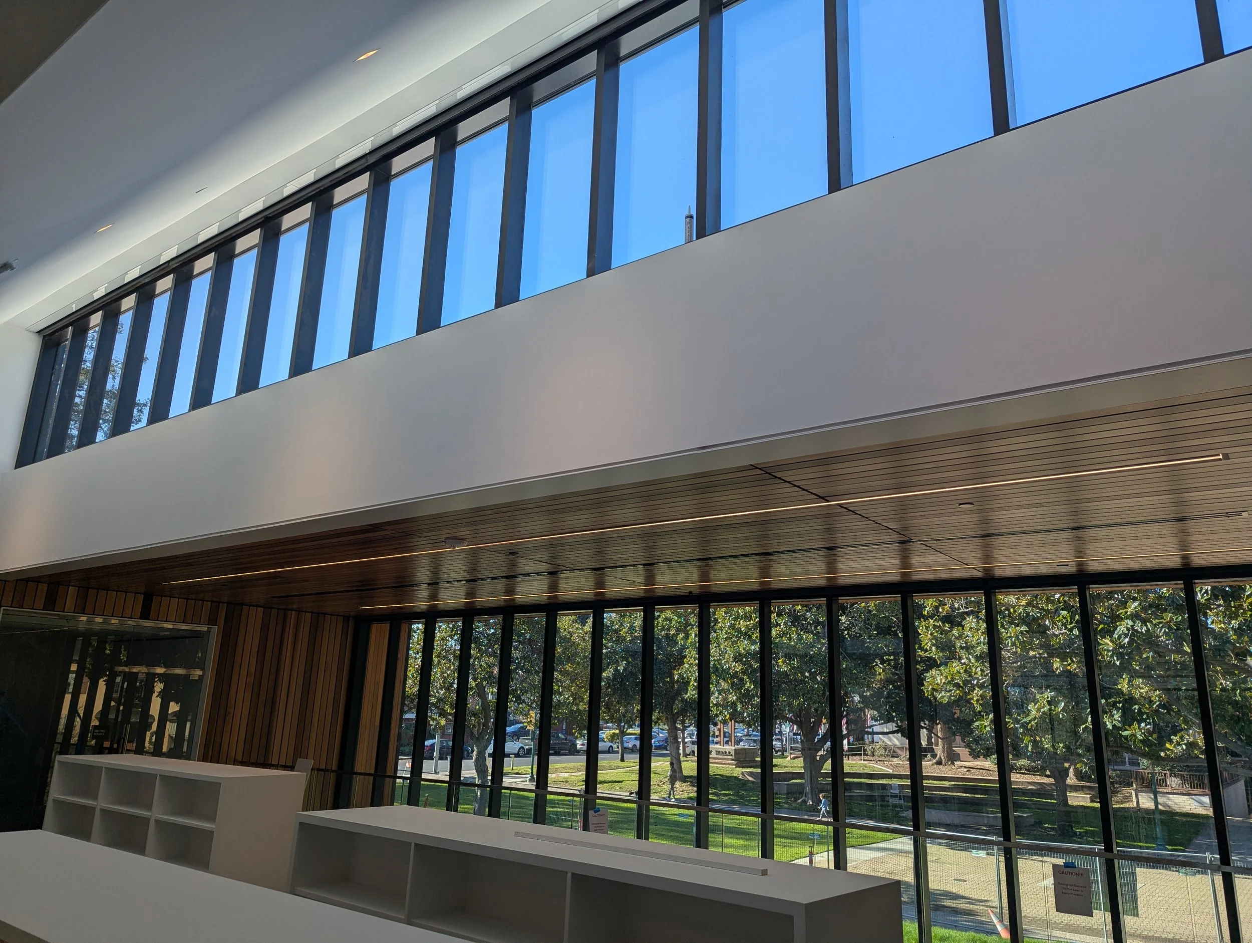 Interior view of the Campbell Library, with large glass windows, a wooden ceiling, and a white wall with horizontal wooden panels, overlooking a green park with trees and a pathway. Structural engineering by BASE Design in San Francisco.