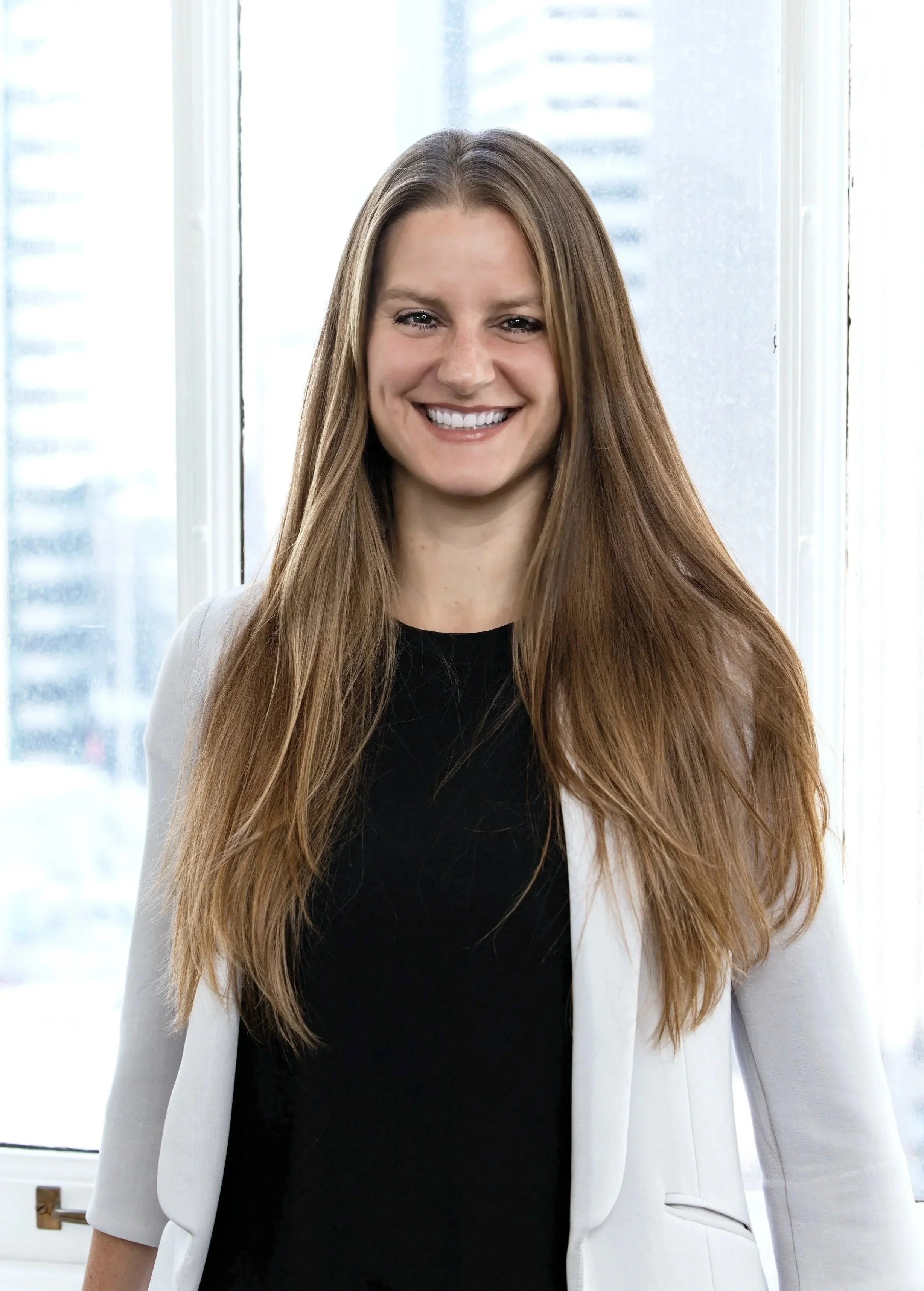 Katy Briggs, Principal and Co-Founder of BASE Design Structural Engineering. She has long brown hair, is smiling, wearing a white blazer and black top, standing indoors near a window with city buildings in the background.
