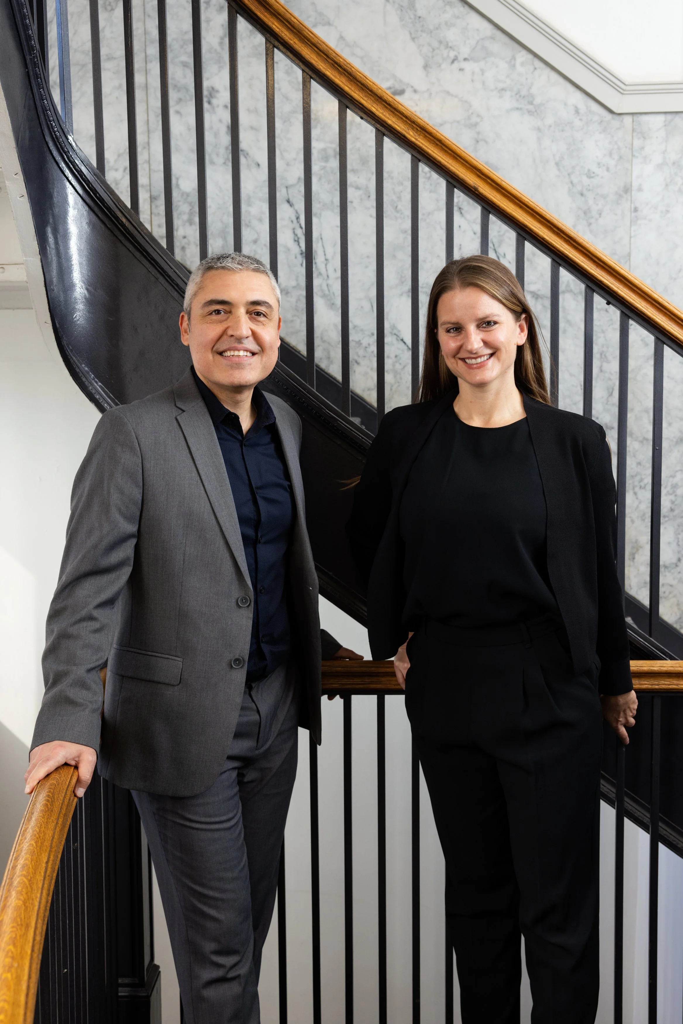 Gokhan Akalan and Katy Briggs, cofounders of BASE Design Structural Engineering in San Francisco, dressed professionally, standing side by side on a staircase with black metal railings and a wooden handrail, smiling at the camera.