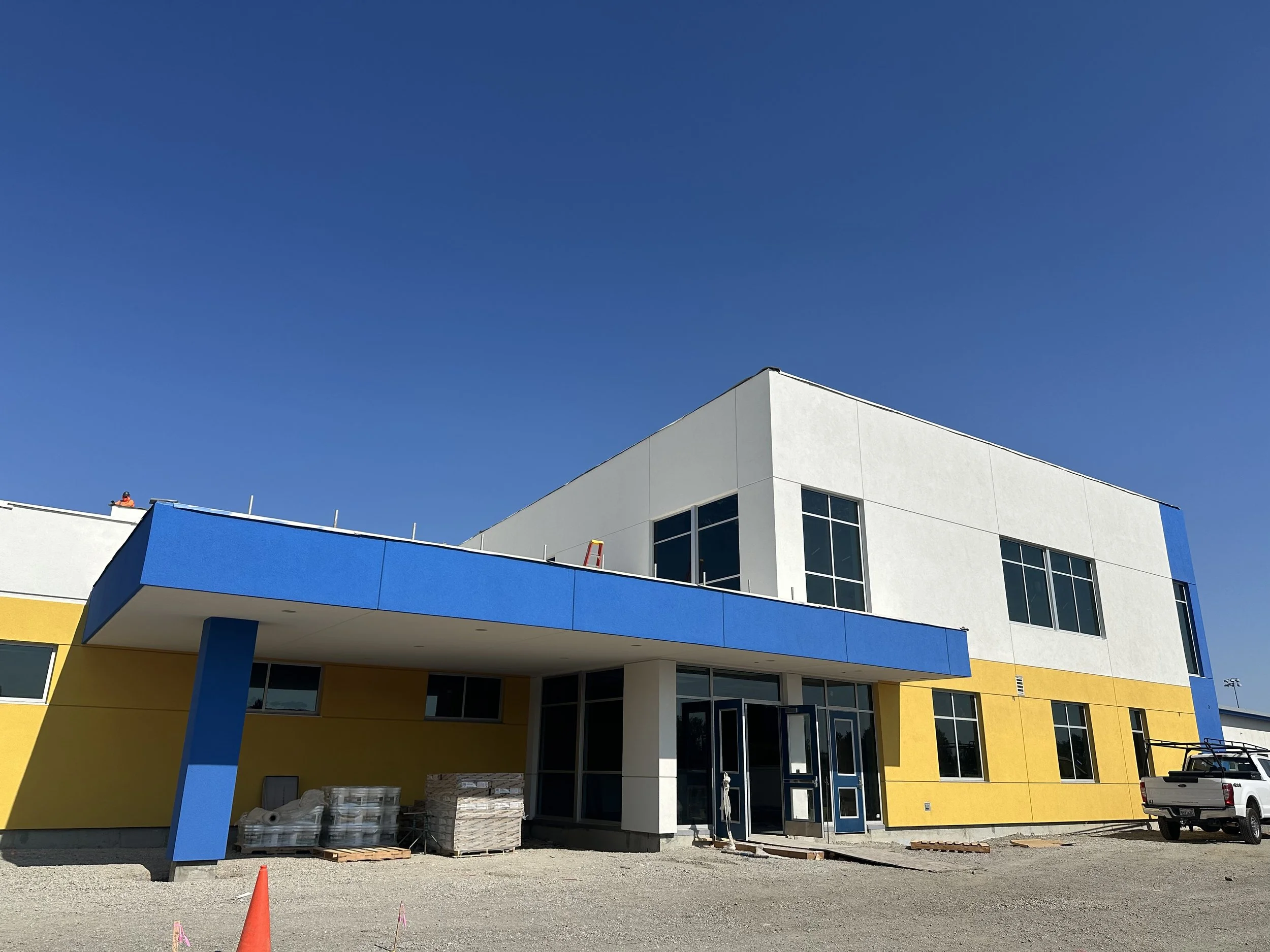 Under construction modern school building with white, yellow, and blue exterior, large windows, and a parking lot in front under a clear blue sky. Structural engineering by BASE Design in San Francisco.