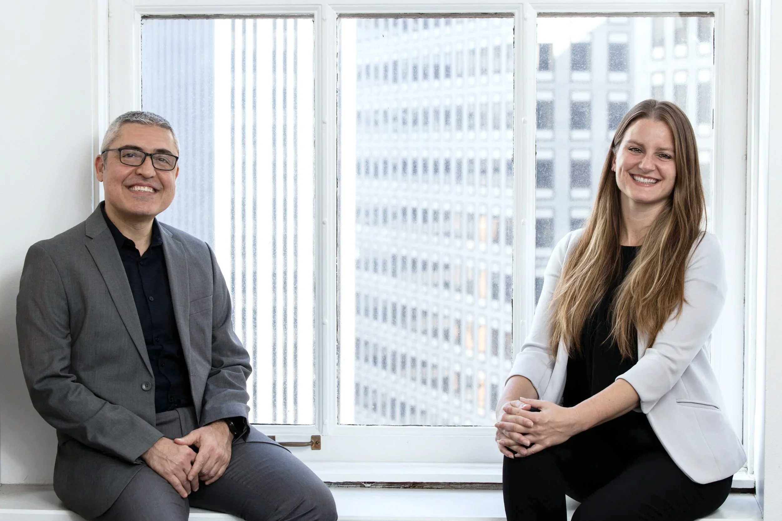 From Left to Right: BASE Design cofounders Gokhan Akalan and Katy Briggs wear informal but professional grey suits, smiling to the camera from their San Francisco office window.