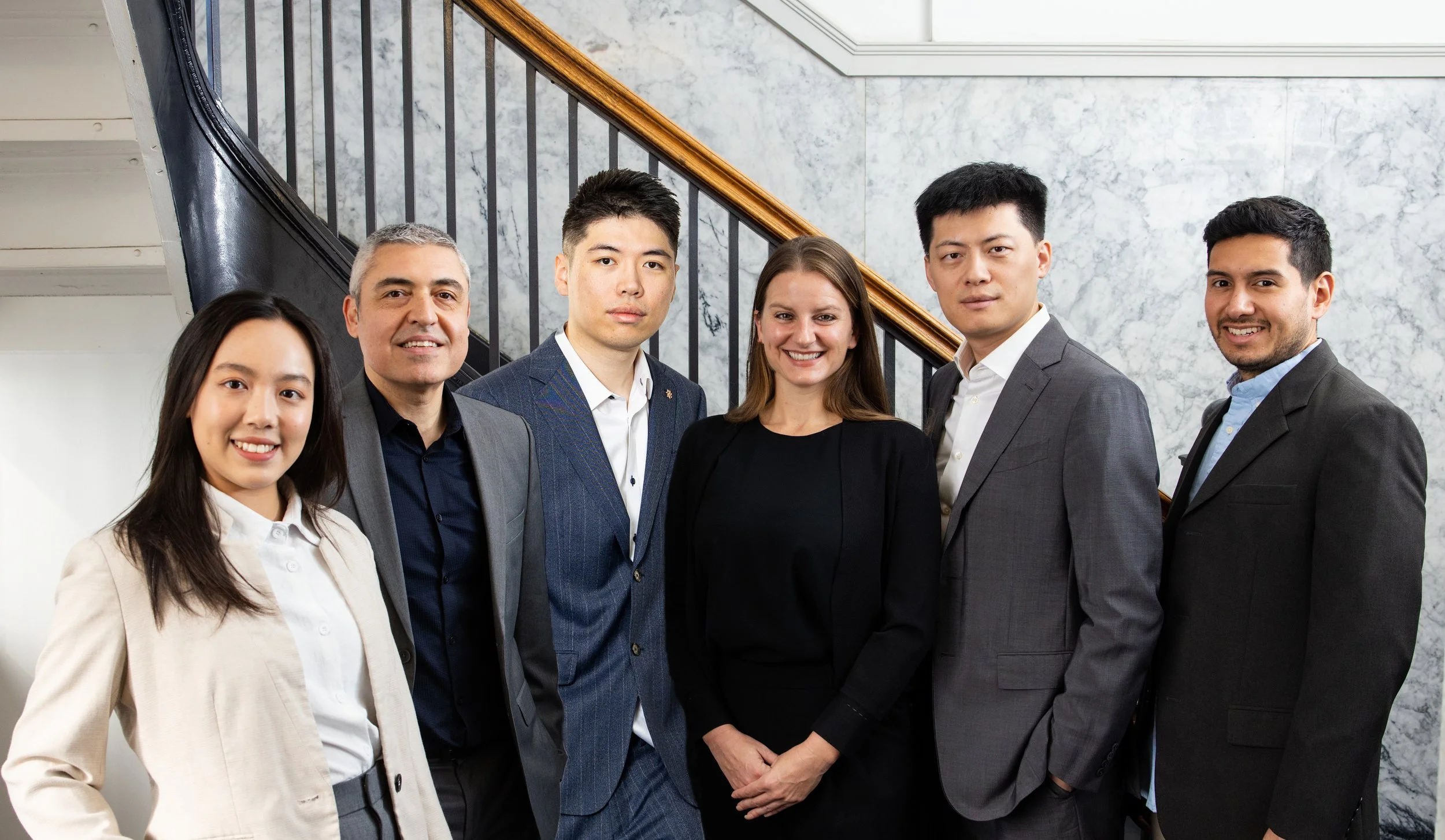 The BASE Design team members, a group of six professionally dressed diverse business people standing in front of a staircase with marble and wood railing at their office building in San Francisco.