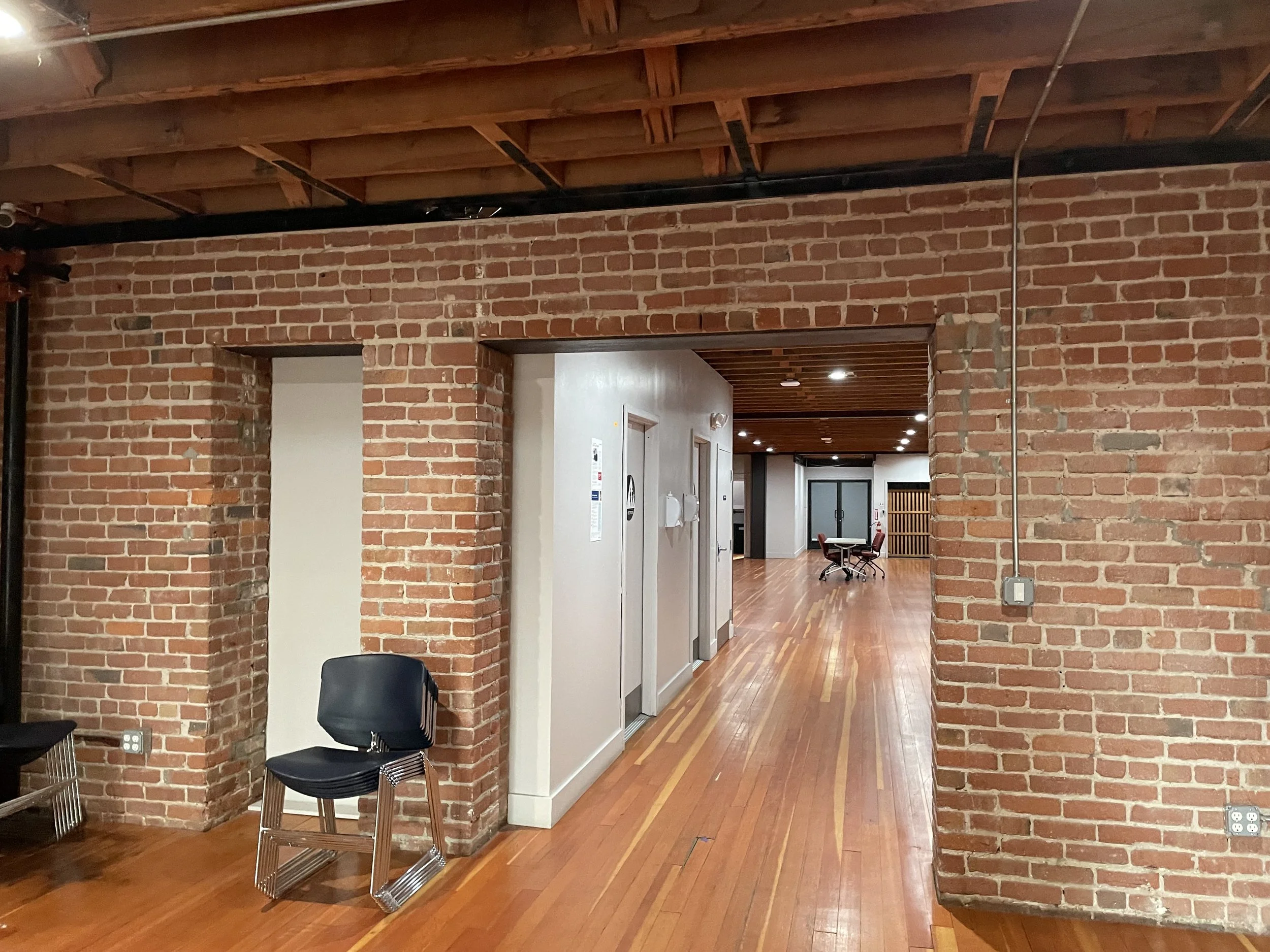 Interior view of a building with exposed brick walls, wooden floors, and a ceiling with wooden beams. An entrance to a hallway leads to a room with seating and tables in the background. Structural engineering by BASE Design in San Francisco.