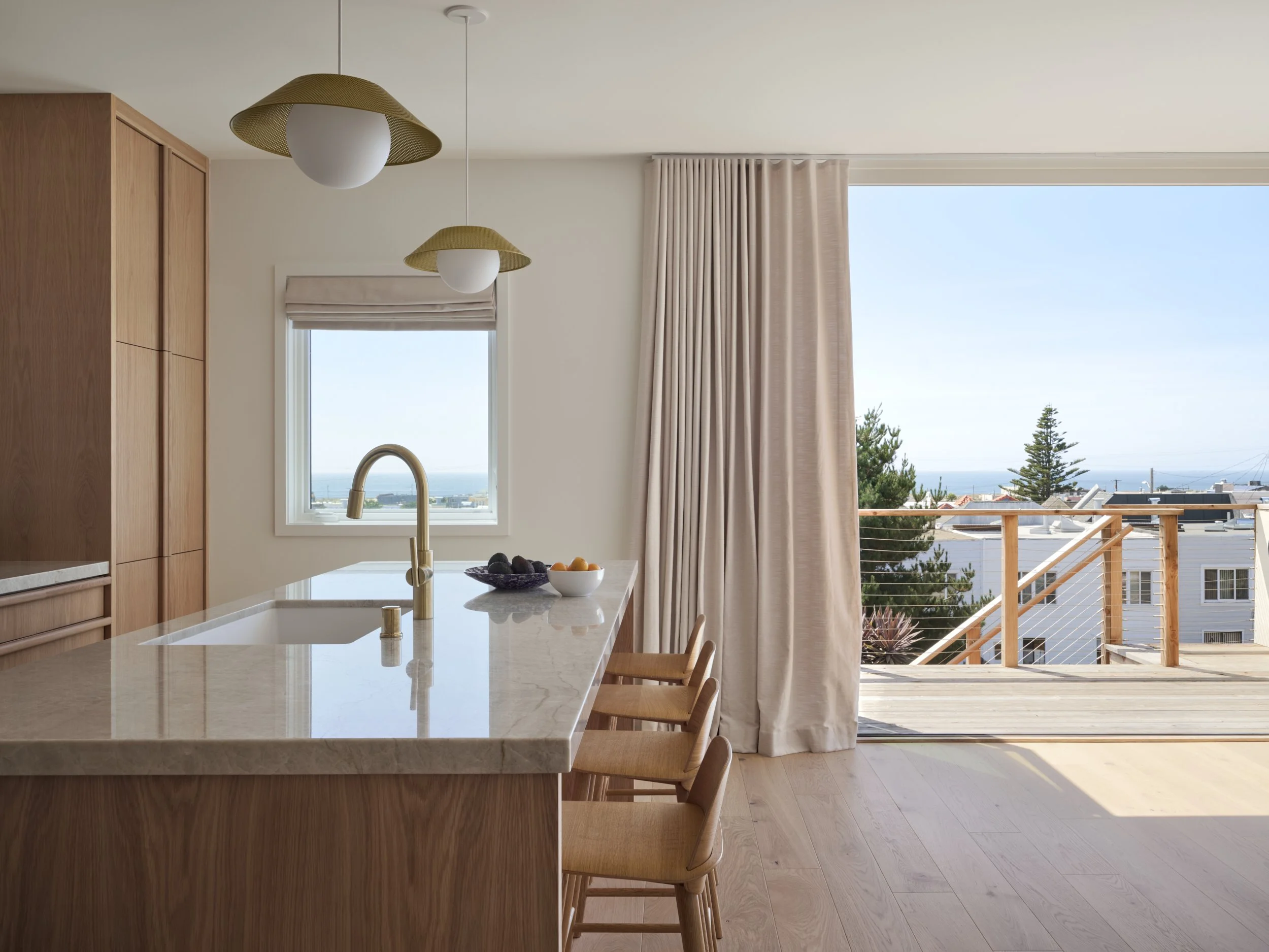 A modern kitchen with a white marble island, a window showing blue sky, and a door leading to an outdoor balcony with a wooden railing, blue sky, and trees. Structural engineering by BASE Design in San Francisco.