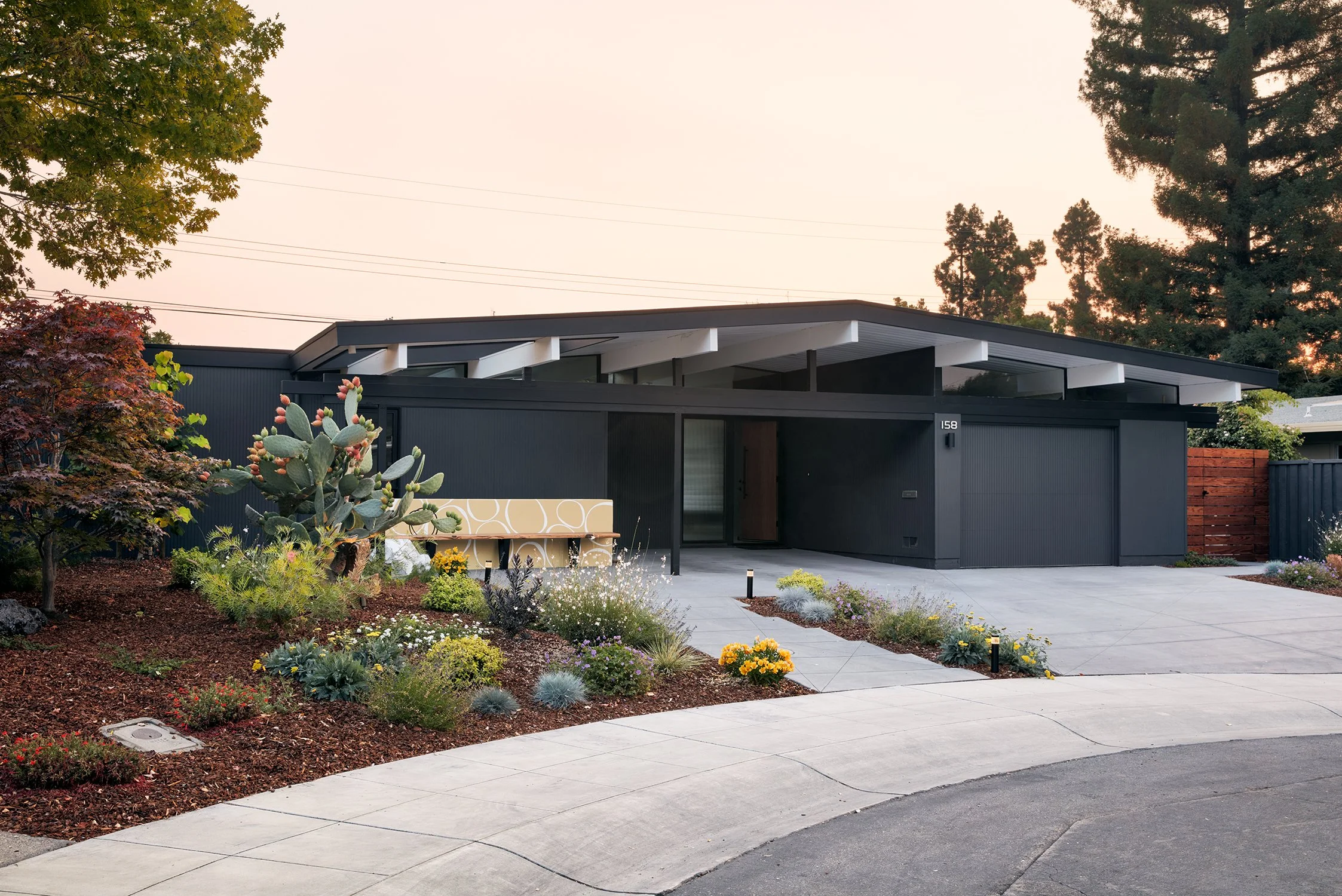 Eichler-desgined home in Palo Alto, CA, with a flat roof, surrounded by a landscaped yard with various plants and flowers, and a light-colored bench near the house entrance.