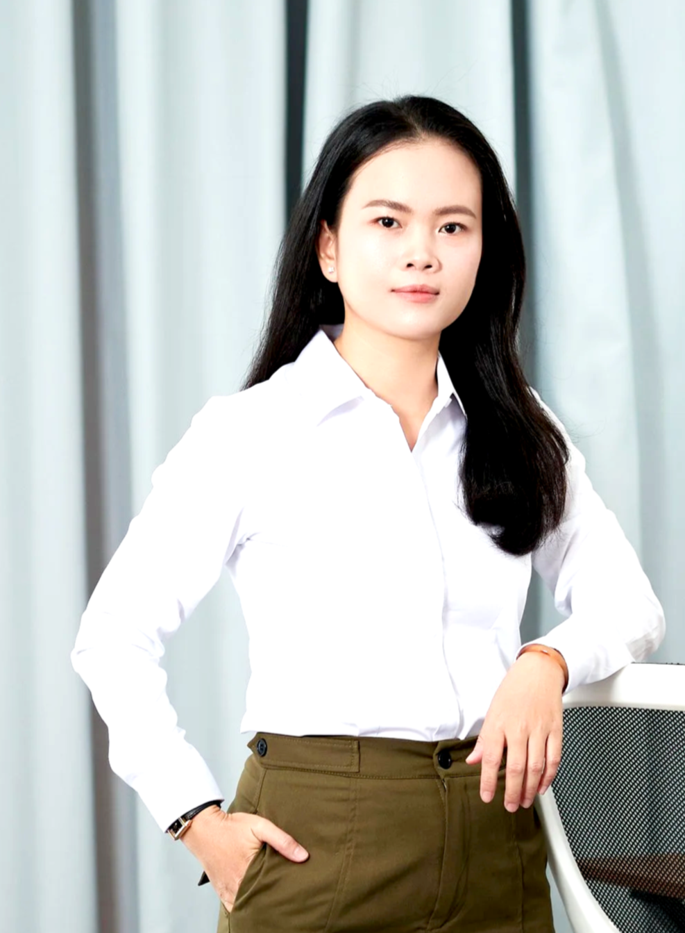 A female member of the BASE Design structural engineering team, with long dark hair, wearing a white button-up shirt and khaki pants, standing confidently in an office with sheer white curtains in the background.