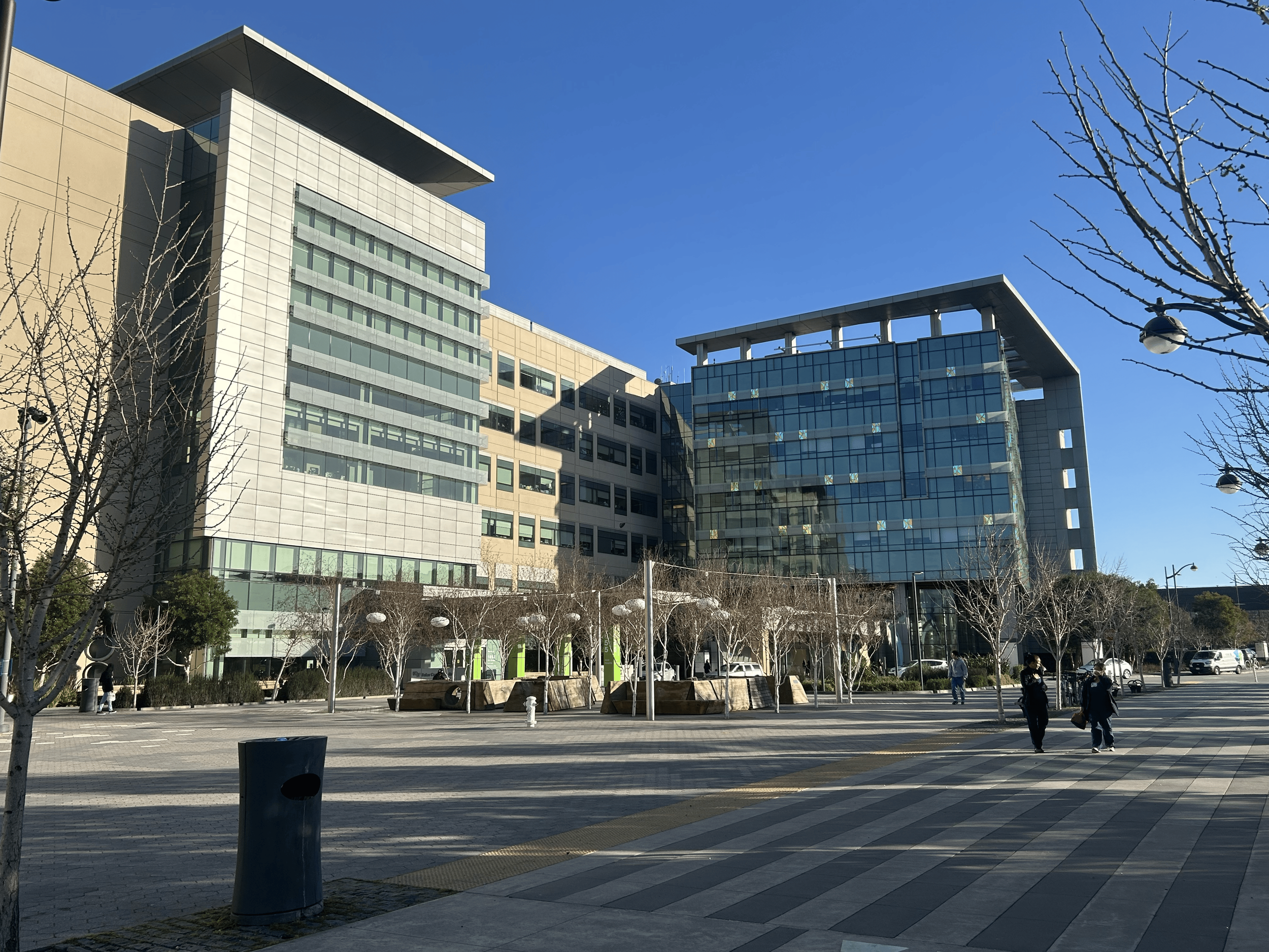UCSF Hospital, a modern office building with glass and concrete facade, trees, and people walking in an outdoor plaza on a clear day. Ongoing structural engineering support by BASE Design in San Francisco.