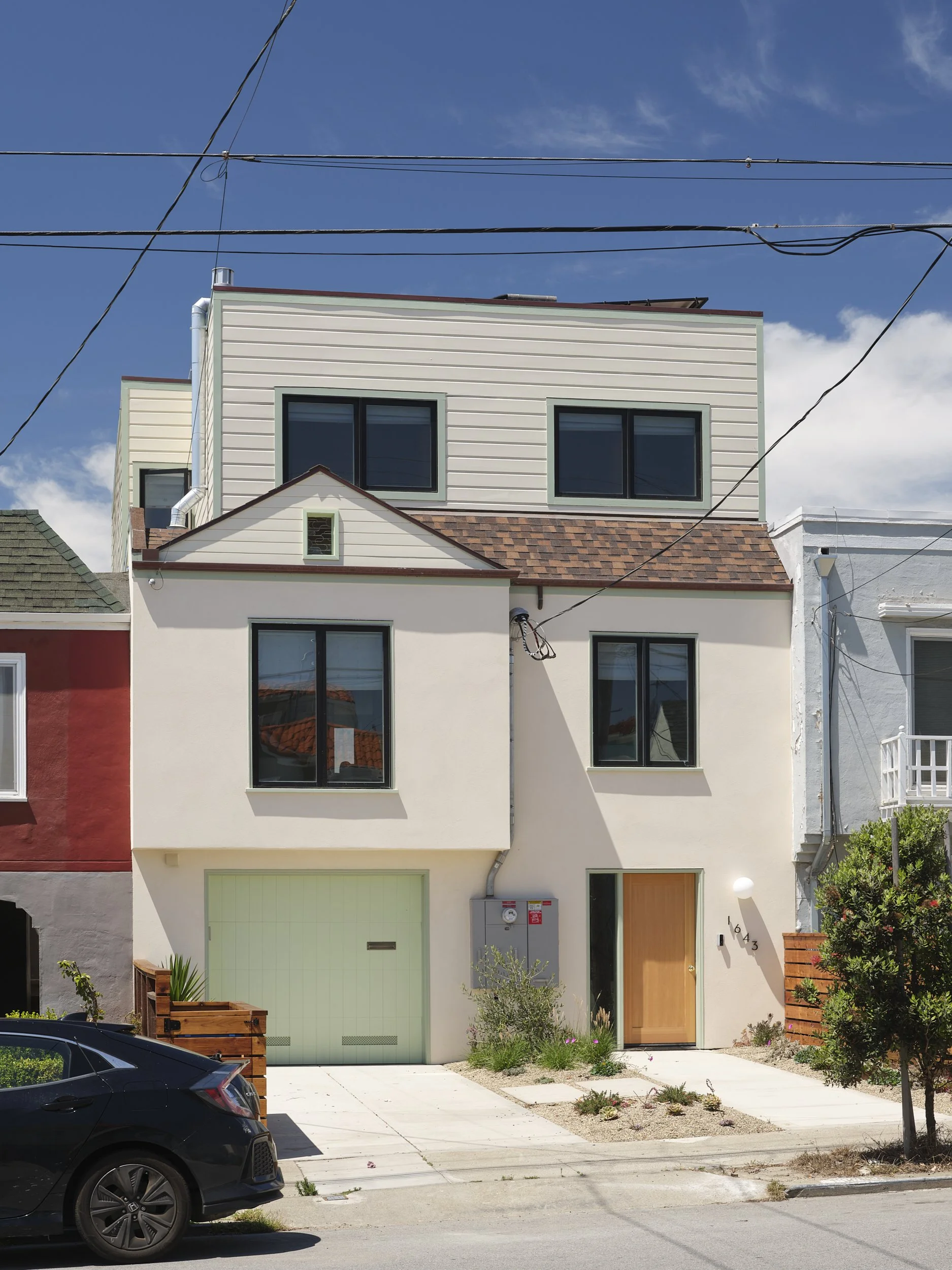 A modern multi-story house with a garage, a front door, and several large windows. The house has beige and white siding, a small front yard with plants, and a black car parked in front. Structural engineering by BASE Design in San Francisco.