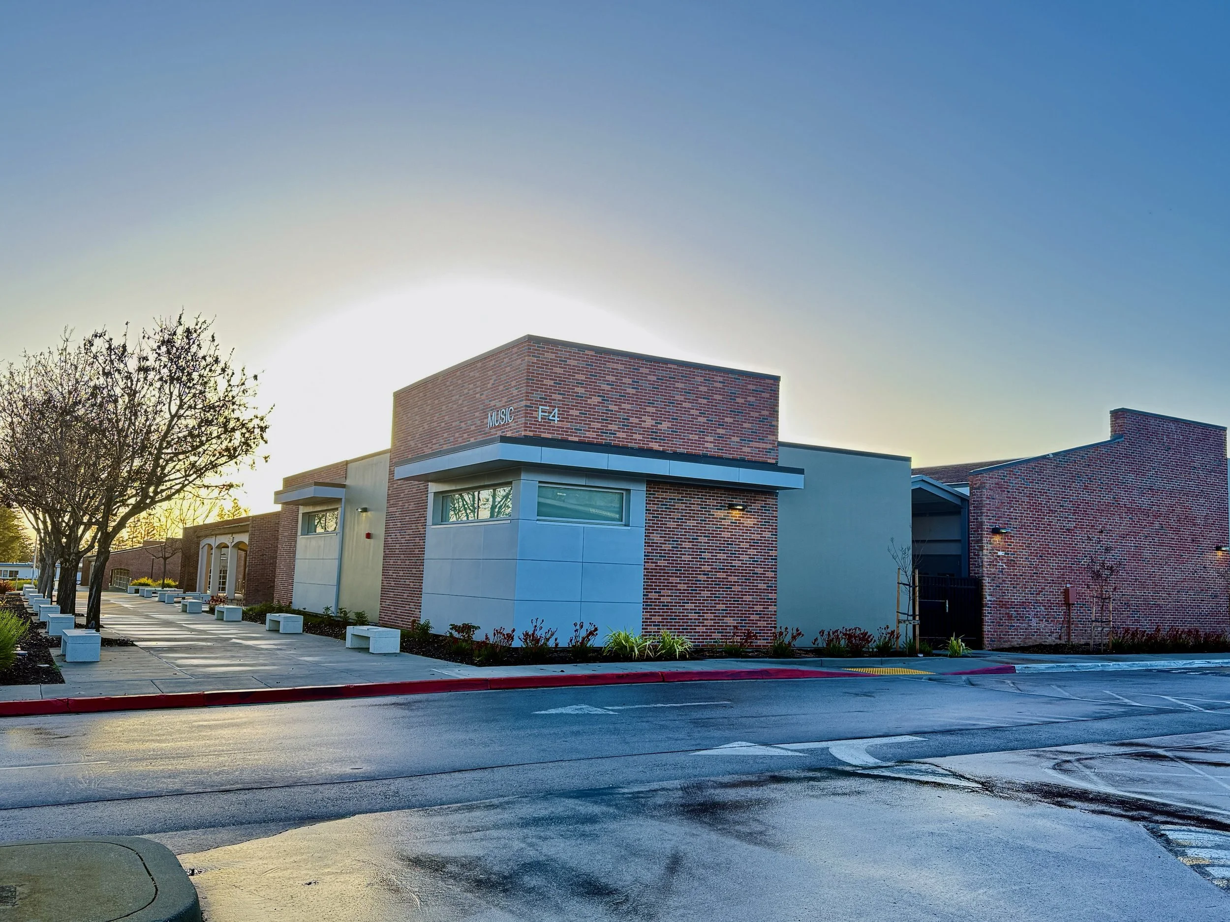 Modern building with brick and concrete exterior, trees lining the sidewalk, and a freshly wet road in front, sun setting behind the building. Structural engineering by BASE Design in San Francisco.